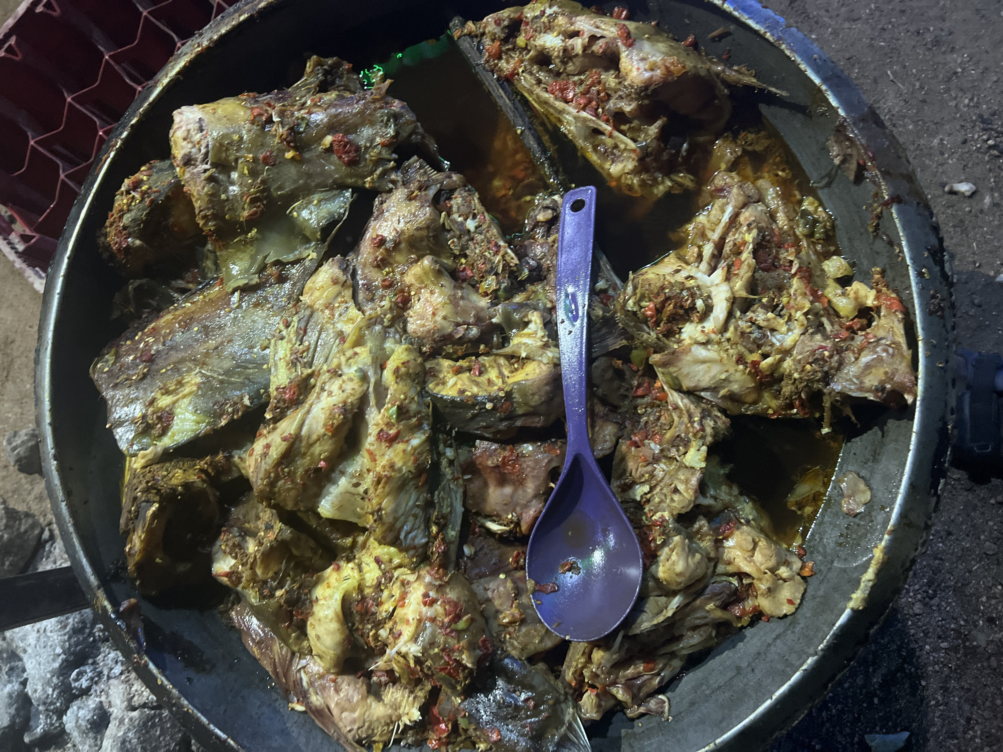 A high-angle shot shows a large, dark-colored metal pan filled with what appears to be cooked fish. The fish is cut into large pieces, seemingly the heads and portions of the bodies, and is seasoned with a reddish spice. A large purple spoon rests on top of the fish. The pan is resting on a rough surface that may be the ground, with patches of dark soil or gravel visible. In the background, glimpses of what appear to be a wire mesh structure and possibly the edge of a vehicle or container are seen. The photograph seems to have been taken at night or in a dimly lit environment, as the lighting is poor. No people are visible in the frame. Based on the prompt, the location would be Jalingo, Nigeria.