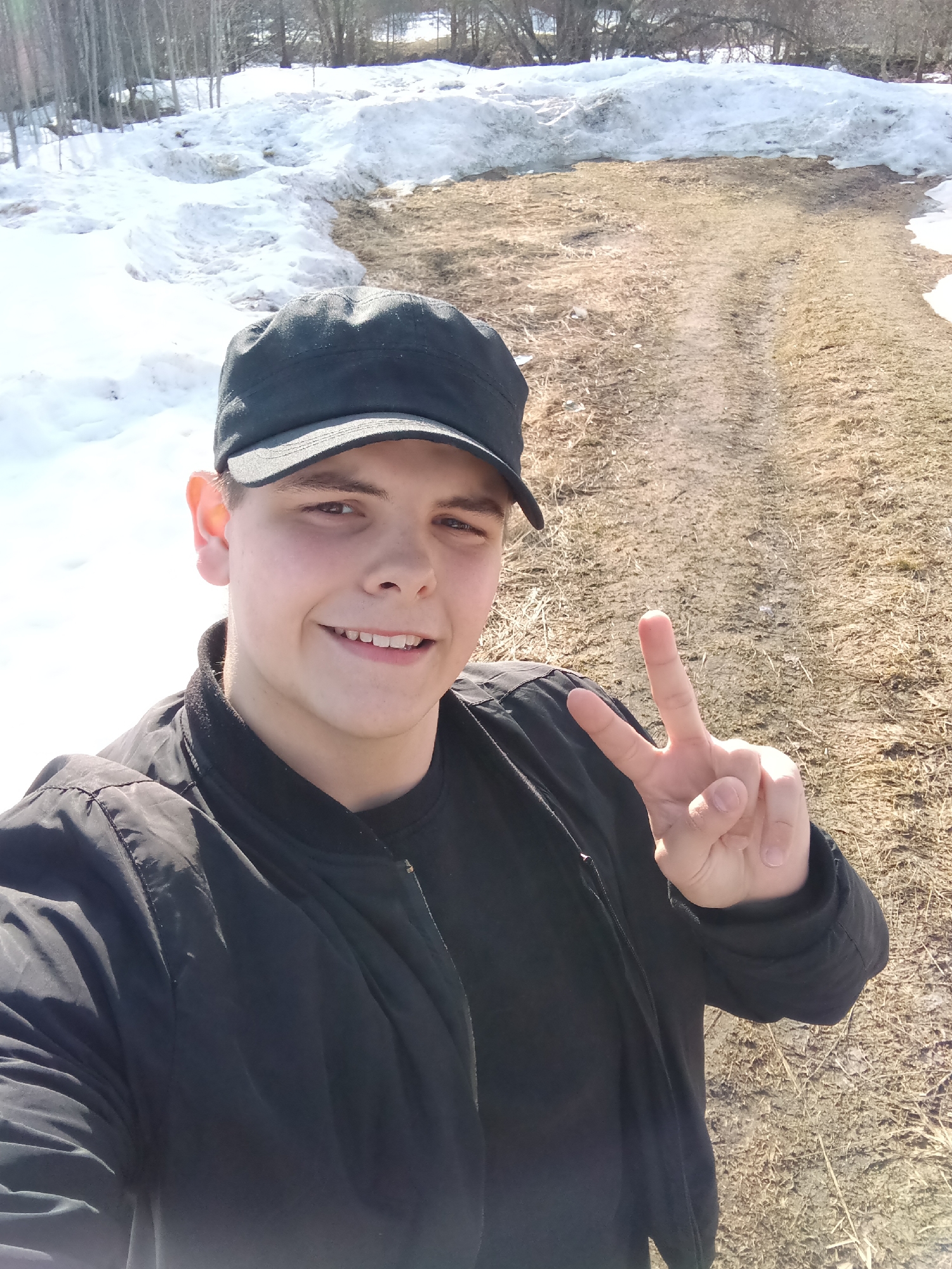 A young adult male, presumed to be in Красный Маяк, Russia, is depicted in a close-up, waist-up selfie outdoors. He is wearing a dark, possibly black, baseball cap, a black bomber-style jacket with a zipper, and a black t-shirt underneath. The male has short, dark hair, brown eyes, and is smiling, revealing his upper teeth. His right hand is raised to chest height, forming a "peace" or "victory" sign with his index and middle fingers extended and separated. The background features a partially thawed landscape, with patches of white snow prominent on the left and a dirt or gravel path with sparse brown vegetation visible on the right. Further in the background, bare deciduous trees are visible under a bright, sunny sky.