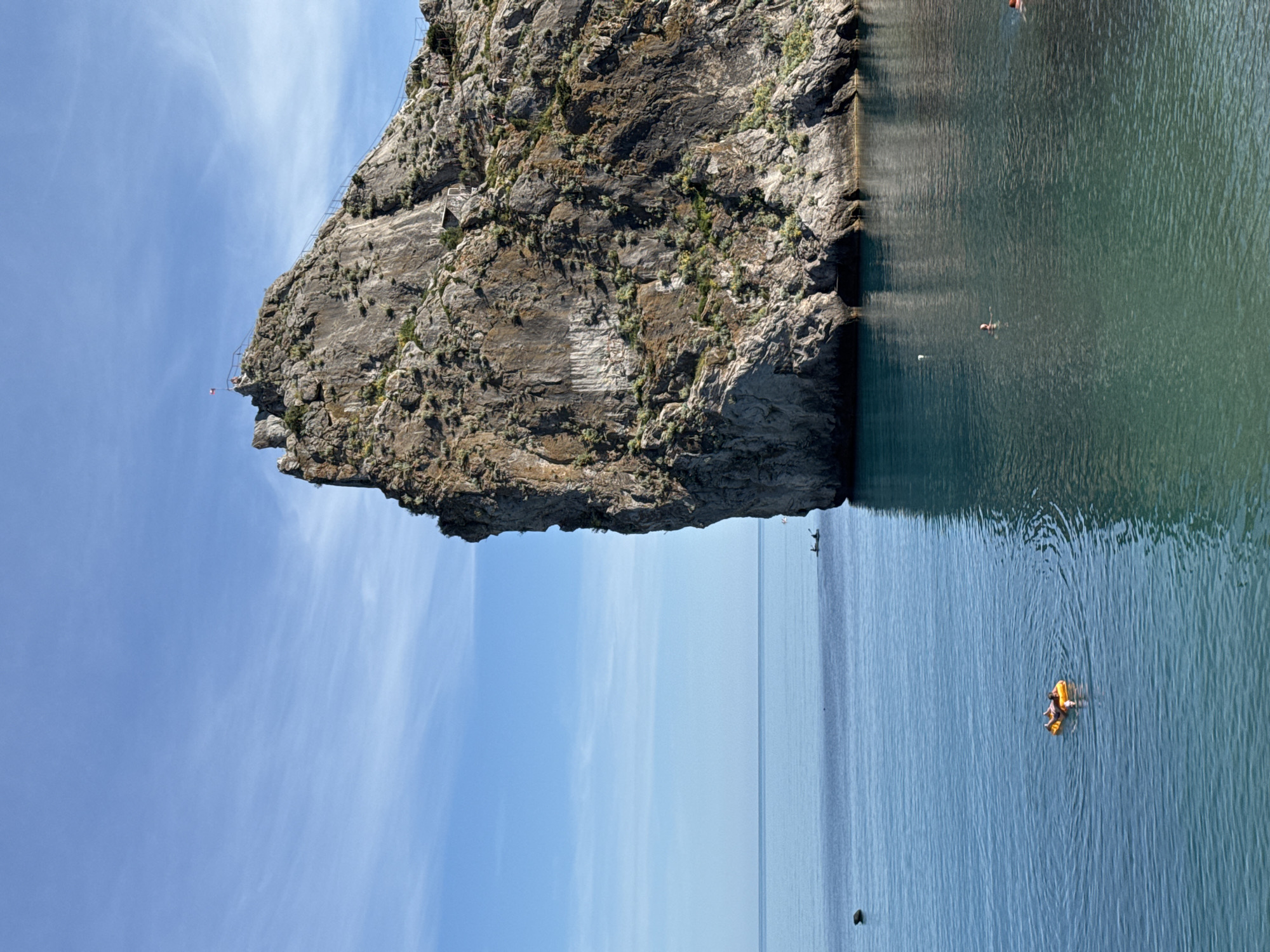 This media file captures a scenic view of a rocky cliff face rising from a calm, blue body of water under a bright sky with wispy clouds, suggesting a sunny day. The cliff itself is rugged and textured, with sparse vegetation growing on its surface. A narrow path or staircase with railings is visible along the cliff's edge, leading to a small platform near the top where a red and white flag is flying.
In the water, there are several individuals engaged in recreational activities. One person is swimming, appearing as a small silhouette in the darker, greener water near the cliff. Further out in the clearer blue water, another person is lounging on a bright yellow inflatable raft. Other faint figures can be seen in the water in the distance. The water's surface is gently rippled, reflecting the sky and the cliff. The overall impression is one of a peaceful summer day at a coastal location, likely a tourist spot given the infrastructure on the cliff and the presence of swimmers and sunbathers. There is no visible text in the image.