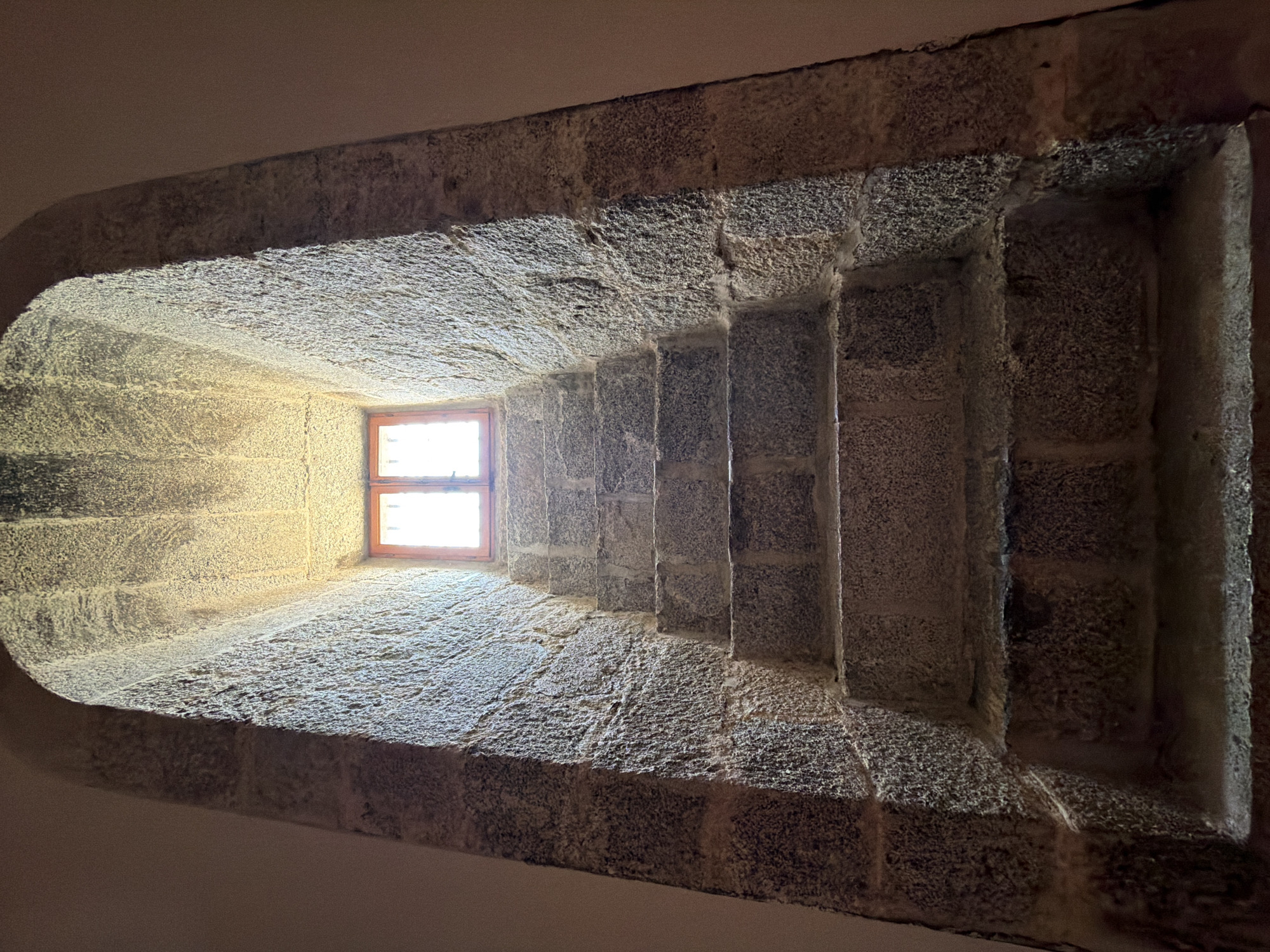 This is a low-angle, full shot of a stone staircase leading up to a small, arched window. The staircase is made of rough-hewn, grey granite blocks, with seven visible steps. The walls surrounding the staircase are also made of similar stone and curve inward to form an arched opening. Sunlight streams through the window, casting a warm glow on the upper portion of the stairs and the ceiling of the arch. The window itself appears to be a simple wooden frame with glass panes, divided into four sections. The environment is dimly lit, suggesting an interior space within a historical building. The image was taken in San Lorenzo De El Escorial, Spain, likely within the Monastery of San Lorenzo de El Escorial, given its architectural style and location. There are no people or animals present in the image.