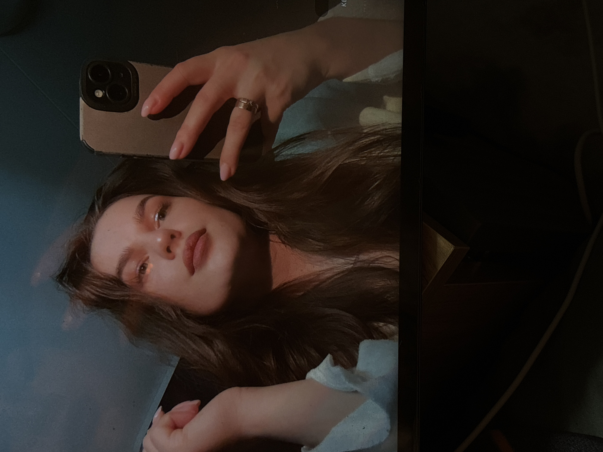 A woman with long brown hair is taking a selfie in what appears to be a dimly lit room. She is holding a smartphone with a gold case in her right hand, and her left hand is visible near her chin. The scene suggests an indoor setting with a dark background, potentially a corner of a room with some furniture visible on the right. There is no information about Chaikovsky, Russia in the image.