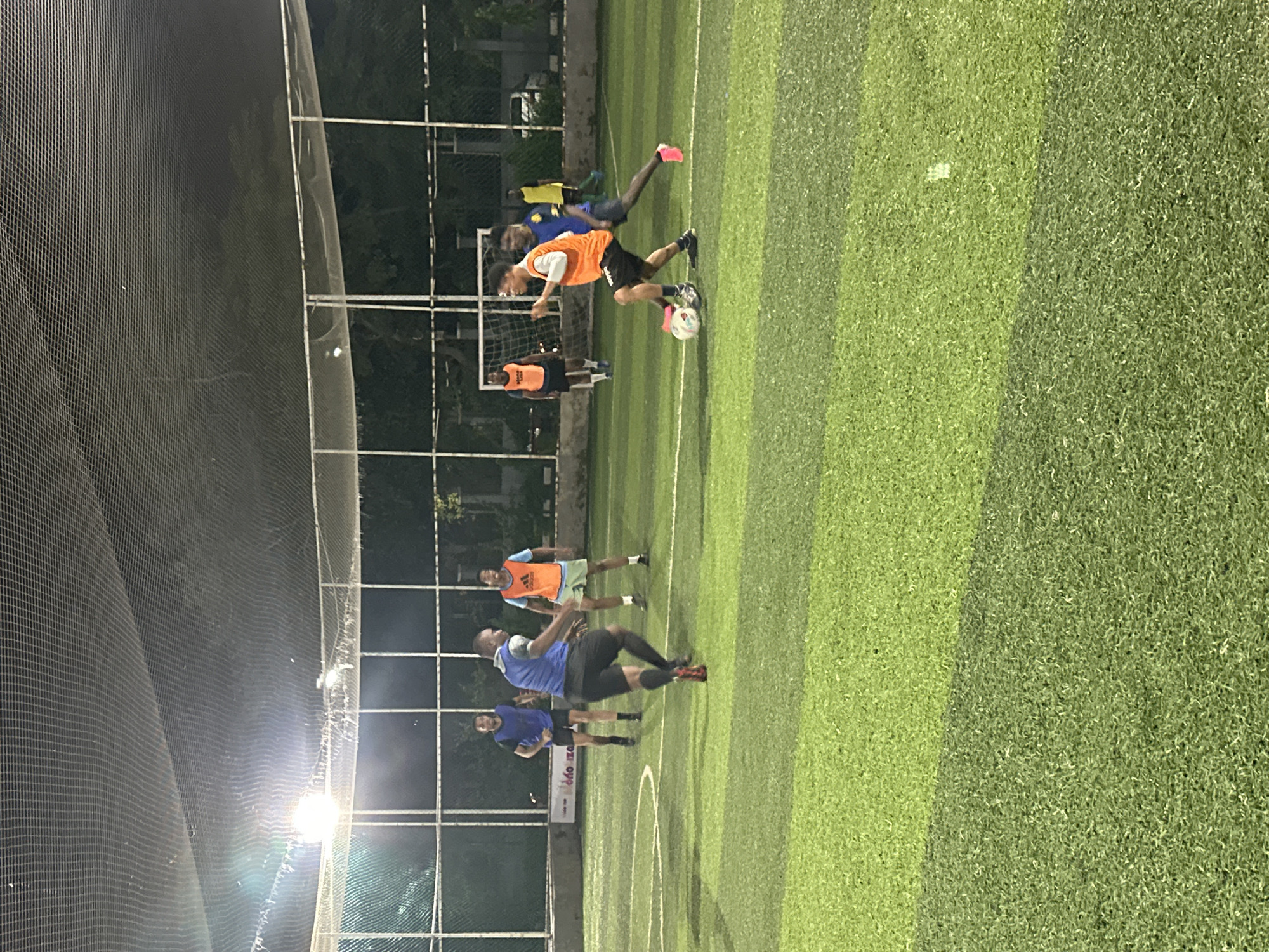 The image depicts an outdoor, nighttime soccer match on a turf field. Several individuals, likely young men, are engaged in play. One player in an orange vest and black shorts is dribbling a soccer ball. Another player in a blue vest and black shorts is positioned behind him, appearing to defend. In the background, other players in similar athletic attire can be seen on the field. The field is illuminated by artificial lights, casting a glow on the green turf and white markings. A net enclosure surrounds the playing area. The scene appears to be set in a sports facility, possibly within the city of Abuja, Nigeria, given the context of the request. The overall atmosphere is one of active recreation and sport.
