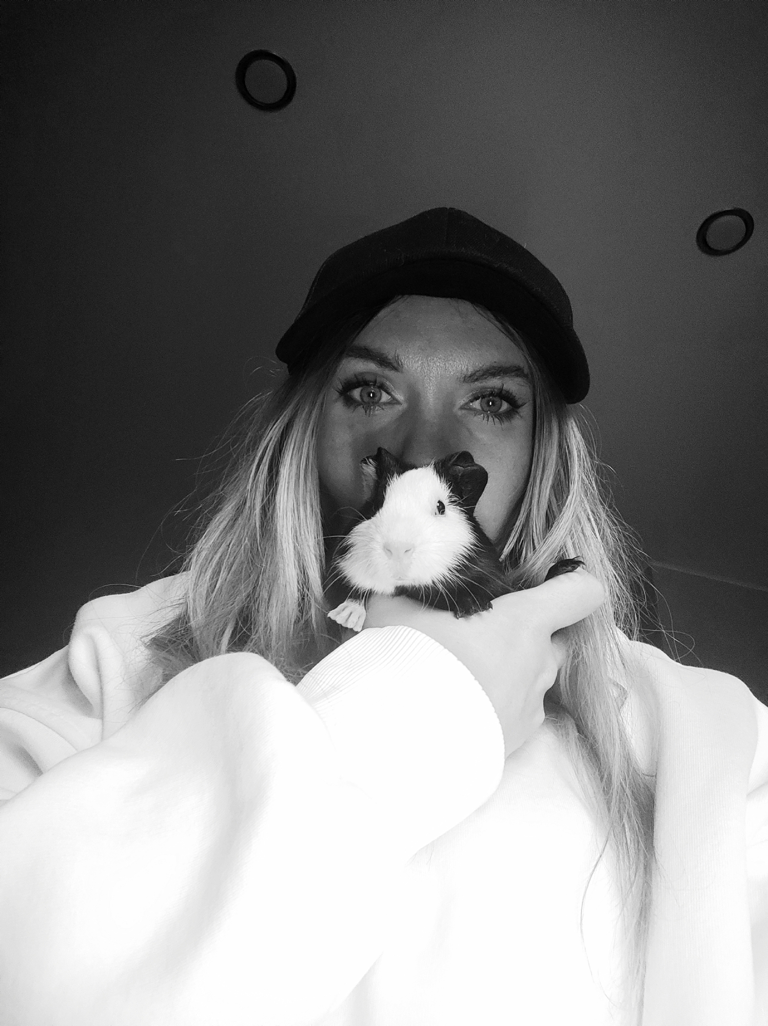 A high-contrast, black and white, low-angle portrait features a person holding a small guinea pig close to their face. The subject, likely female, wears a dark baseball cap and a light-colored (appearing white) hoodie or sweatshirt, with long, light hair visible cascading over their shoulders. Their eyes are prominent and directly engage the camera, offering a neutral to serious expression. The guinea pig, with distinctive black and white fur, is held in the person's hand, positioned in front of their mouth and nose area, and also appears to be looking towards the camera.
The background is dark and plain, suggesting an indoor setting. Two circular objects, possibly light fixtures or vents, are visible on the dark surface above the person's head. The image is devoid of any specific time of day or weather indications due to its indoor setting and black and white presentation. No text is visible. While the provided context indicates Votkinsk, Russia, there are no visual cues within the image itself to confirm this location.