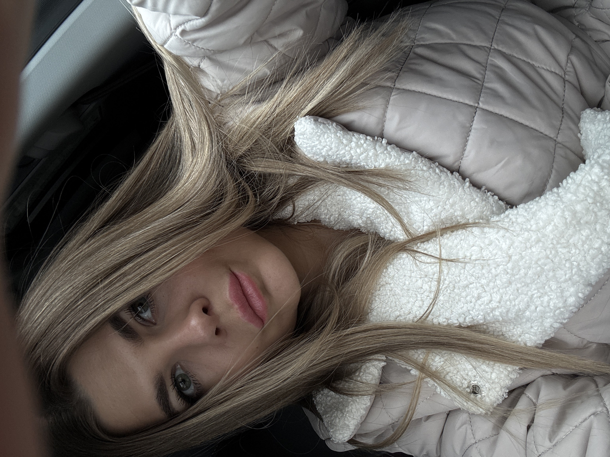 The image depicts a female individual in a close-up, angled portrait, situated within the interior of what appears to be a vehicle. She has long, straight, light blonde or brown hair, green eyes, and prominent eyebrows. Her lips are colored with a pink hue. She is wearing a light beige, quilted jacket featuring a white, textured, fleece-like lining around the collar and lapel area, with a silver snap button visible on the right lapel. The background is dark and indistinct, consistent with vehicle interior panels. A partial view of a hand or finger is visible in the upper left corner, suggesting the image is a selfie. No external landmarks or environmental features are present to determine a specific location within the city of Chaikovsky, Russia.