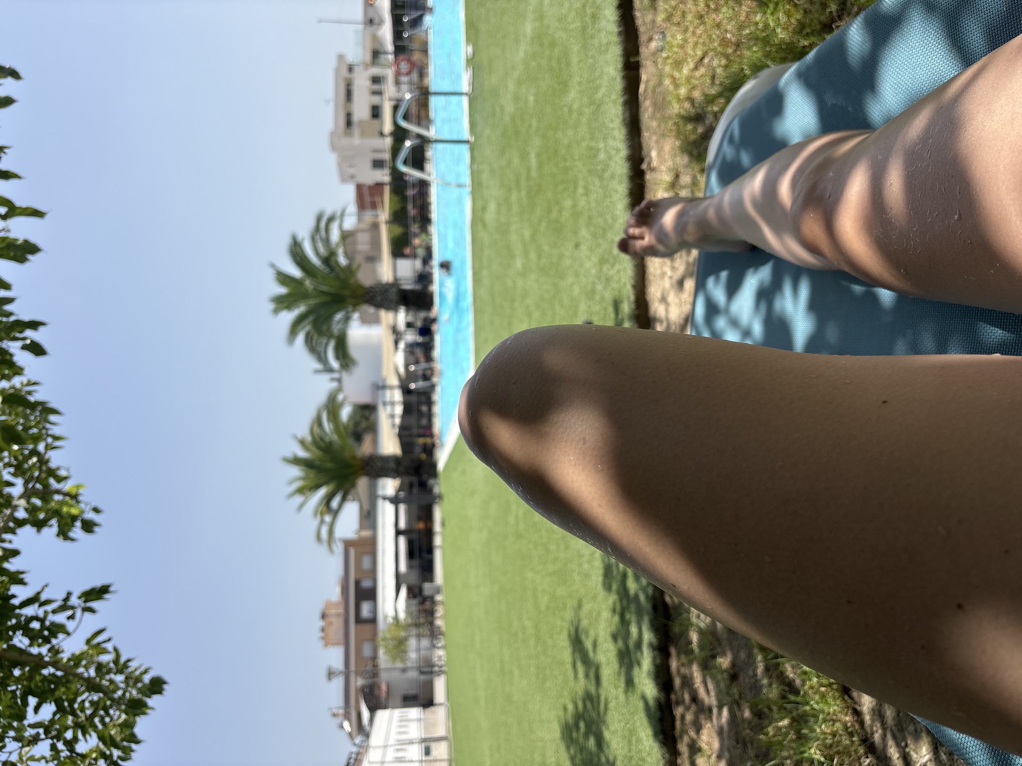 The image shows a person lying on a blue lounge chair on a sunny day. The person's legs are prominently featured in the foreground, with one leg in sharp focus and the other slightly blurred. The legs appear to be slightly damp, suggesting they may have recently been in water. The lounge chair is positioned on a patch of grass, with dappled sunlight filtering through overhead tree branches.
In the background, a swimming pool with bright blue water is visible. Several people are in the pool, some swimming, and others standing near the edges. Palm trees are scattered throughout the scene, and buildings, likely part of a resort or residential area, can be seen beyond the pool. The sky is clear and blue.
The location is identified as the city of Cox, Spain. The overall setting is a recreational area, possibly a public pool or a resort, offering a leisure experience under the sun.
