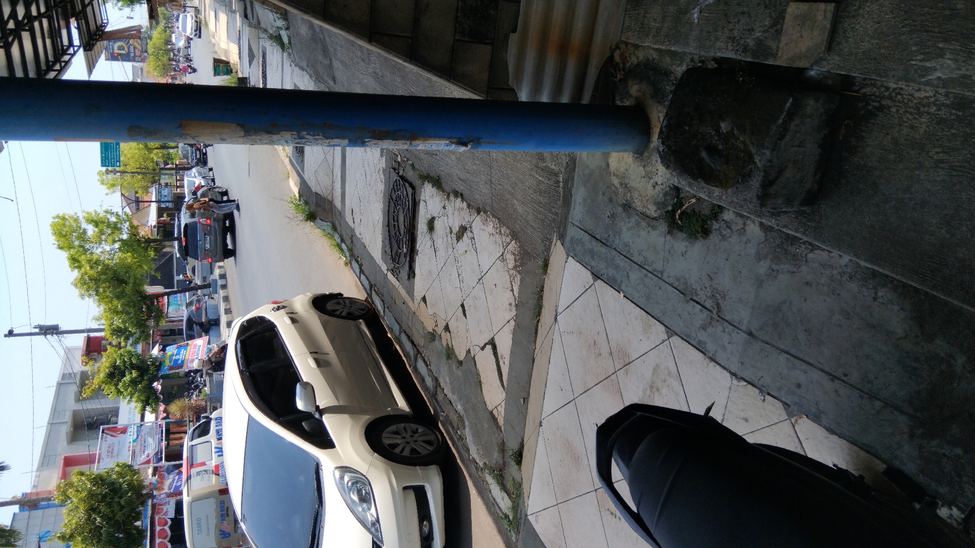 The image shows a street scene in Batang, Indonesia, on a sunny day. A white hatchback car is parked on the left side of the street, facing away from the viewer. The car is a Honda Jazz. In the background, there are buildings, trees, and other cars on the road. A blue pole is visible in the foreground on the right side of the image, with a weathered texture and some peeling stickers. The street has a concrete sidewalk with cracked tiles and patches of grass growing through. A metal manhole cover is visible on the sidewalk. Further down the street, several people are walking and riding motorcycles. The scene suggests a typical urban environment in Indonesia with its characteristic architecture, vehicles, and vegetation.