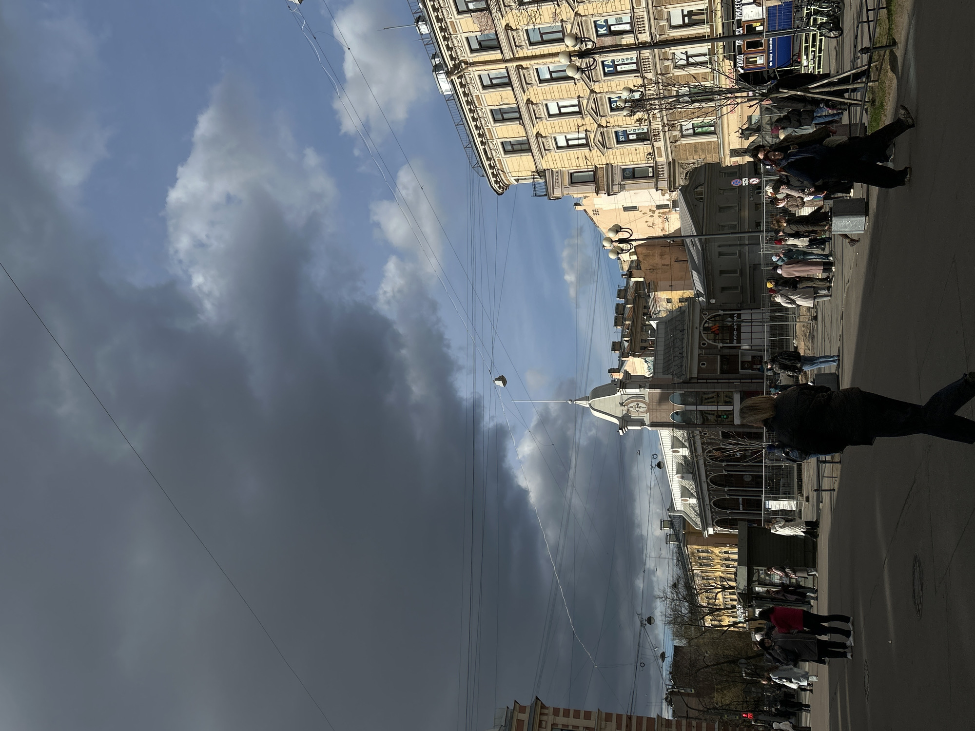 The image captures a vibrant street scene in Saint Petersburg, Russia, under a dramatically contrasting sky. The perspective is slightly upward, focusing on the blend of architectural details, street activity, and the dynamic weather.
On the right side of the frame, a row of ornate, multi-story buildings with classical yellow-beige facades lines the street. These buildings feature numerous windows, decorative elements, and ground-level storefronts. A complex network of overhead wires, likely for electric public transport like trams or trolleybuses, crisscrosses the sky above the street.
The sky itself is a notable subject, divided between clear blue sky with some scattered white clouds on the far right and a large expanse of heavy, dark grey storm clouds dominating the left and central portions of the frame. This sharp contrast suggests rapidly changing weather conditions.
On the wide sidewalk and street below, several people are visible, walking and standing. They appear dressed in jackets and coats, indicating cool weather. Shadows cast on the ground suggest that sunlight, despite the dark clouds, is illuminating parts of the scene, likely from the right. In the midground, a dark statue of a person is prominent, and further back, more pedestrians are seen.
Visible text on building signs includes the Cyrillic word "ВХОД" (Entrance) on a blue sign, and "ПСИХО-МЕТРА" (Psycho-Meter) near a red traffic light on a lower building to the left. Some signs also display Japanese characters. Other discernible but less clear signs may indicate a "BEAUTY SALON" or "FLOWERS" in Cyrillic. The scene depicts a bustling daytime atmosphere in a historic urban setting.
