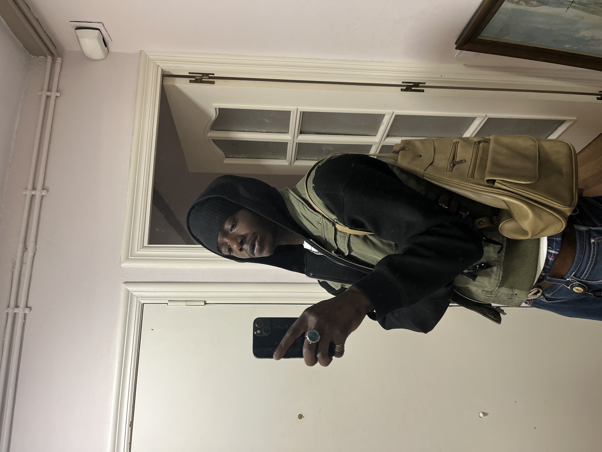 The image is a selfie taken in front of a mirror. The person is wearing a black hoodie, a green vest, and a black beanie. They are also wearing a backpack and have a ring on their finger. The background is a white door with a frame, a white wall, a heating tube, a white ceiling light, and a partially-viewable framed painting in the upper right corner. There is no evidence to suggest this image was taken in Barcelona, Spain.