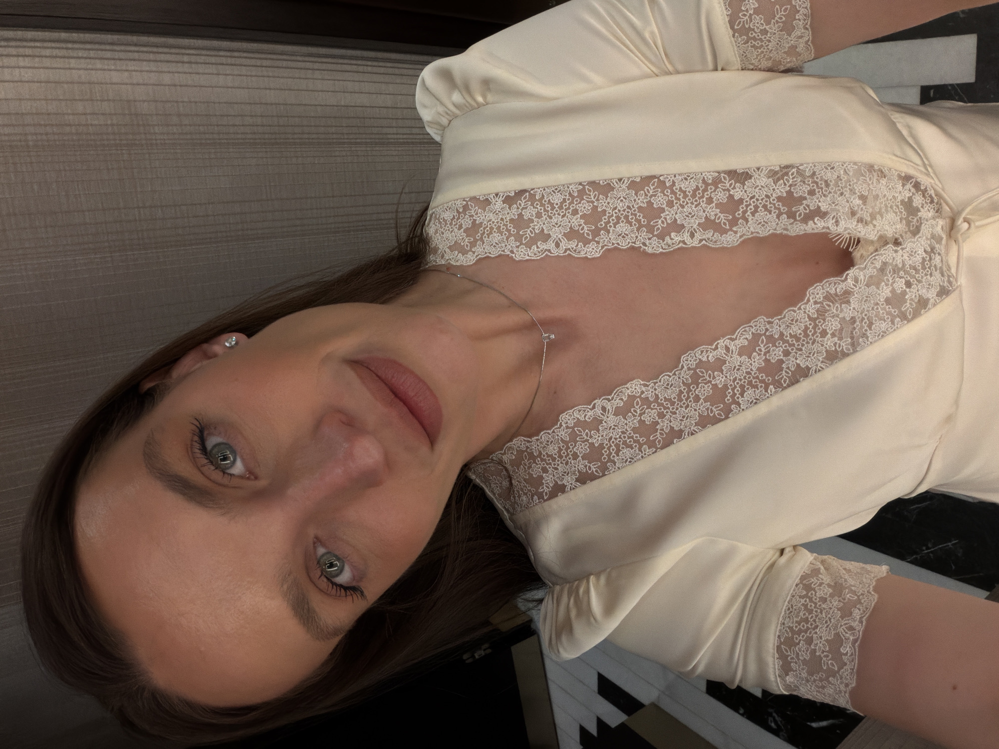 A woman is posing for a close-up selfie while inside. She is wearing a light-colored satin robe or dress trimmed with white lace. The top of a dark wooden cabinet or molding is visible in the background, along with black and white geometric tiles. The woman has brown hair and light eyes. She has a small stud earring in her left ear and a delicate necklace. There is no evidence to indicate that the picture was taken in Budva, Montenegro.