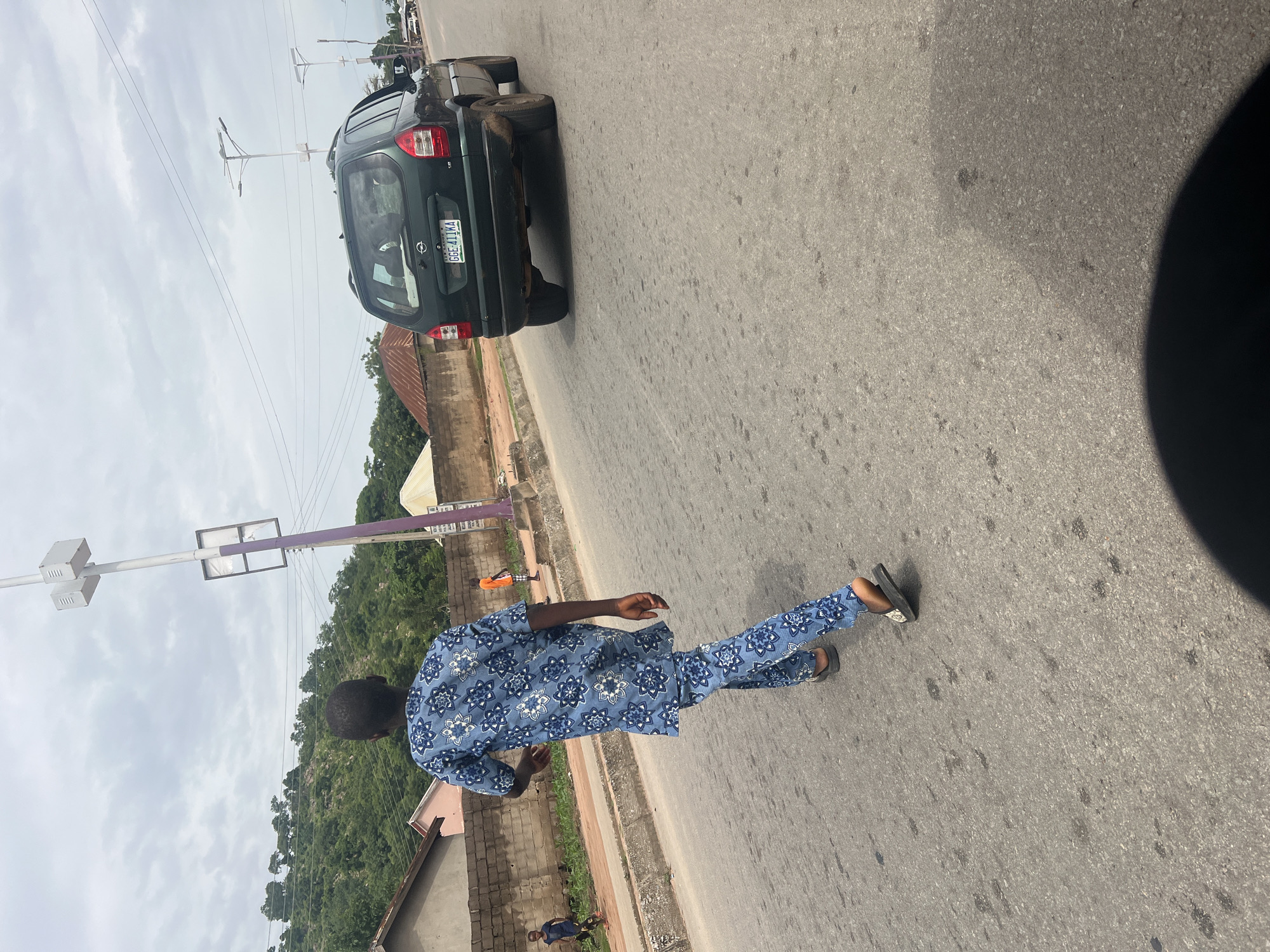 A man in a blue patterned traditional outfit walks on a paved road in Minda, Nigeria. He is wearing dark sandals. In the background, a dark green SUV is parked on the side of the road, its license plate clearly visible. The license plate reads "GGE 411KA". Further in the background, a lush green hillside is covered in trees, with some buildings and utility poles visible. The sky is overcast with clouds. The scene appears to be during the daytime. The man's posture suggests he is walking casually. The road is bordered by a low brick wall and some greenery. Another person in an orange shirt is visible further down the road, near the parked SUV. A third person, a young boy in a dark shirt, is seen standing on the side of the road in the foreground. The overall setting is a roadside in a town or rural area, with natural landscape in the distance.