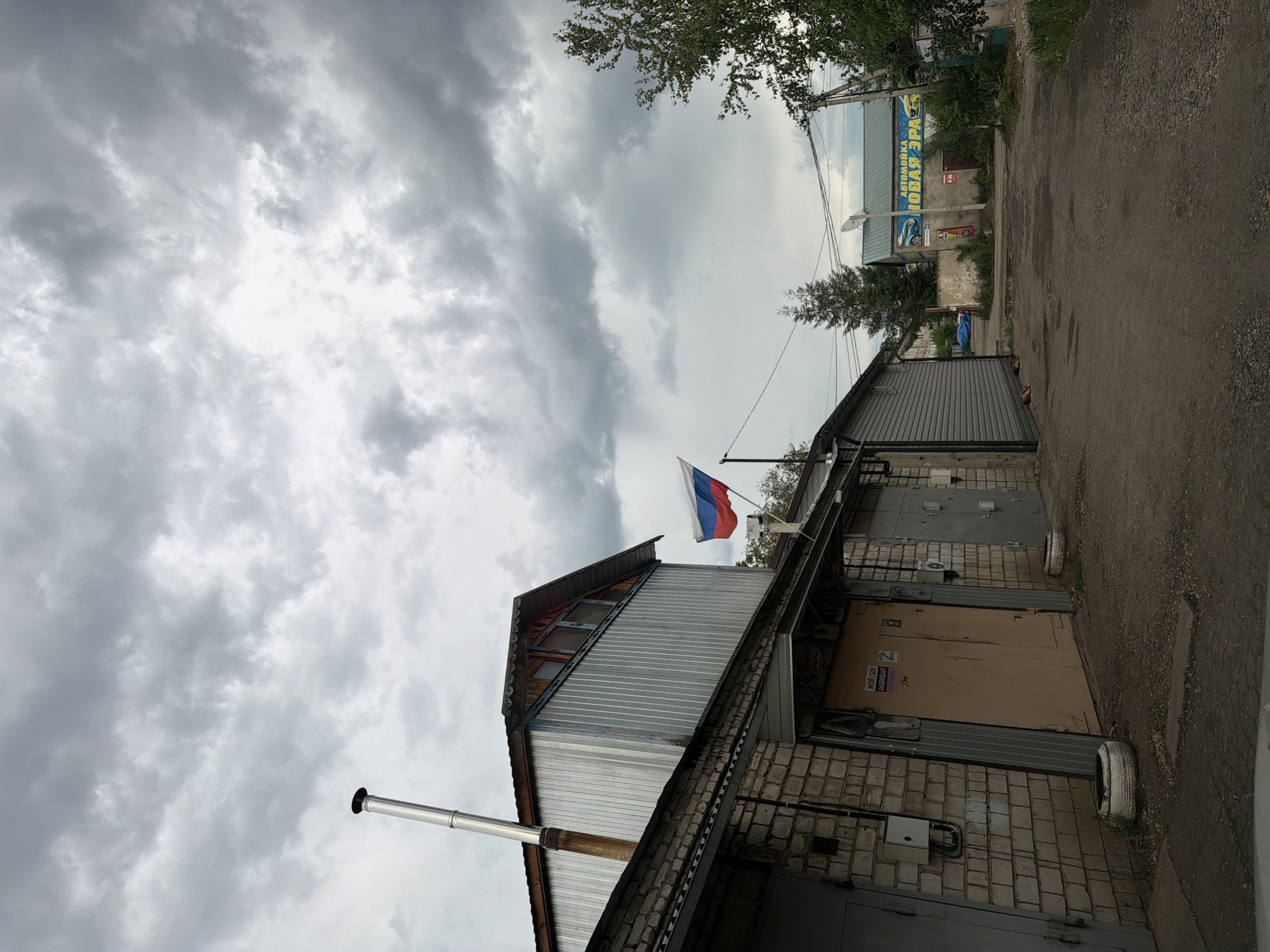 The image depicts a view of a street in Birobidzhan, Russia, with several buildings lining the right side of the road. The sky is heavily overcast with dark clouds. In the foreground, a grey gravel road leads into the distance.
On the left side of the road, there is a long, low building constructed with pale brick, featuring multiple garage doors. One of these doors is a prominent light brown color, and another is a darker grey. A tall, silver metal chimney extends upwards from the roof of a taller section of this building, which is clad in corrugated metal. A Russian flag is visible flying on a pole between two of the garage doors.
Further down the road on the right, there is another building with a green roof and a large blue and yellow sign that reads "АВТОМОЙКА НОВАЯ ЭРА" (Car Wash New Era). A white satellite dish is visible on the roof of a building behind this one. Vegetation, including trees and overgrown grass, is present along the sides of the road and around the buildings, suggesting a somewhat unkempt or rural urban environment. Utility poles and wires are also visible. No people are present in the image.