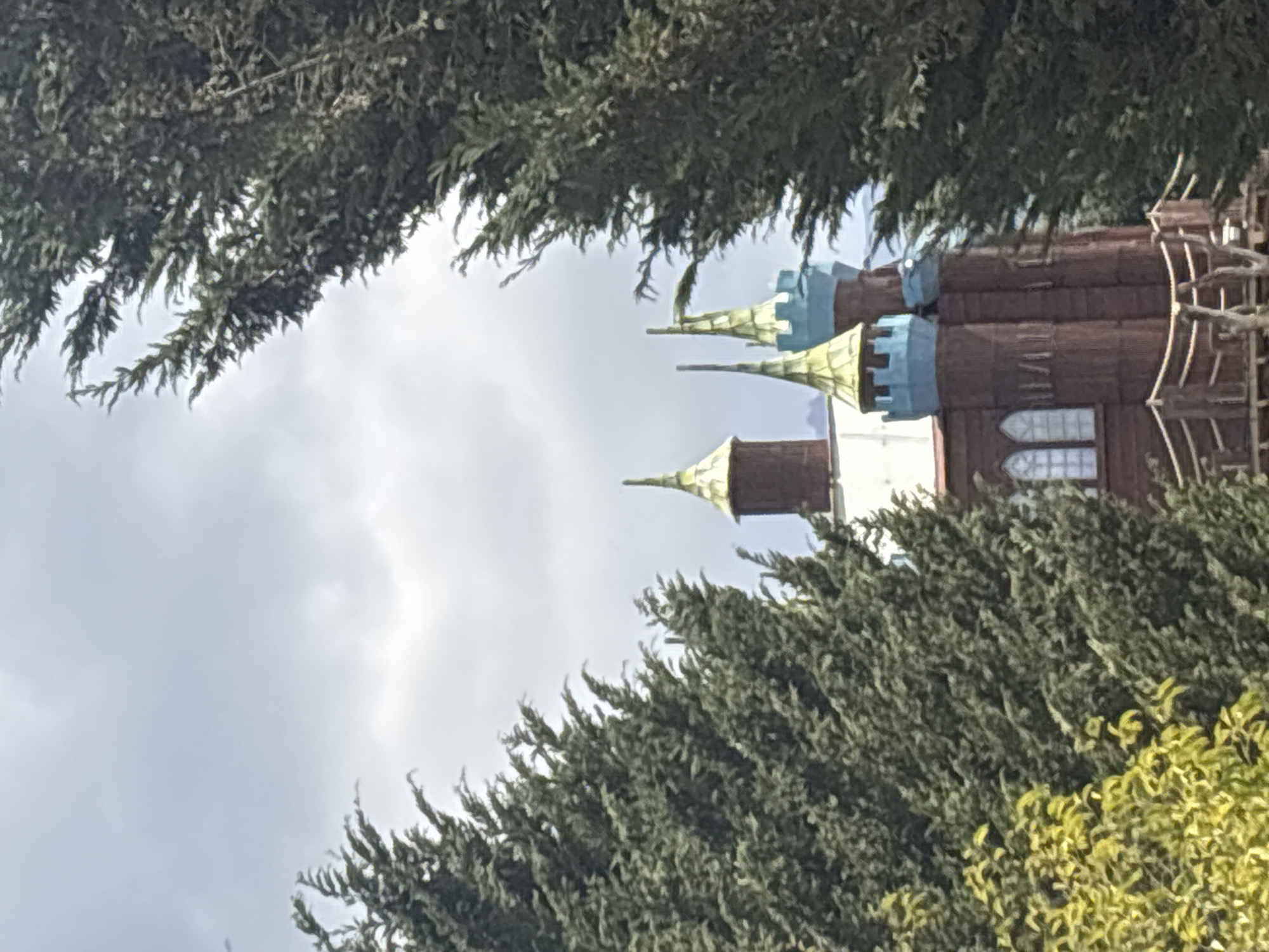 The image shows a building with multiple towers, likely a castle-like structure, with brown and light-blue colors, viewed through the foliage of trees. It is likely located in San Juan Arcos Ojo de Agua, Mexico. The sky is cloudy.