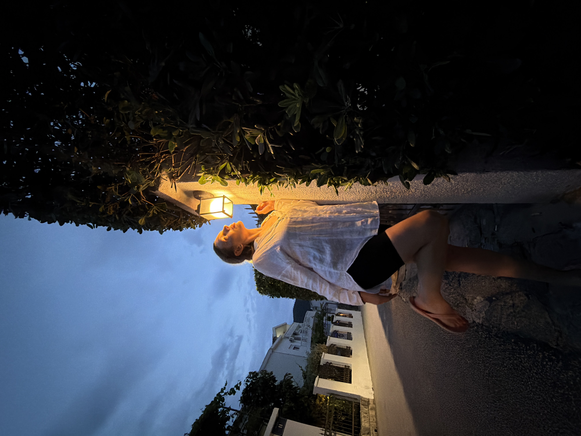 A woman in a white shirt and black shorts stands on a stone wall next to a building in Budva, Montenegro. She is illuminated by a wall-mounted lantern, casting a warm glow on her face and upper body. Her right leg is bent with her foot resting on the wall, and her left leg is extended behind her. The scene takes place at dusk, with a dusky blue sky and silhouetted trees in the background. A street and houses are visible on the left side of the image. The wall is textured and light-colored, contrasting with the dark foliage of the surrounding bushes.