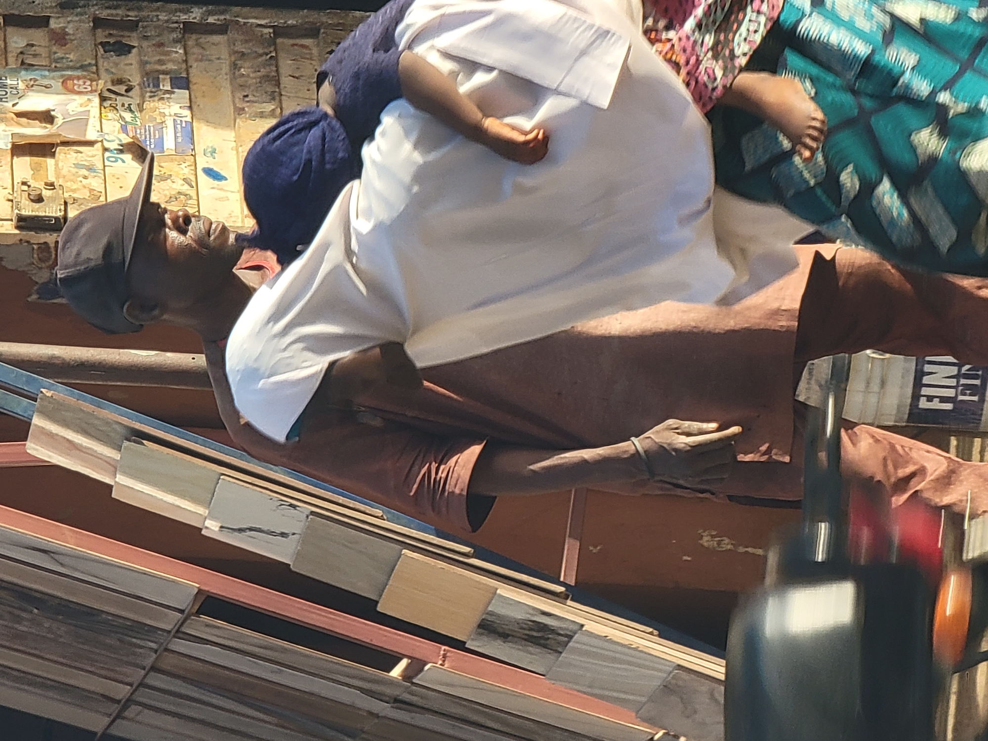 The image, taken in Maiburiji, Nigeria, depicts a street scene in bright daylight. The main subject is a man, positioned upright as if riding a vehicle, wearing a black baseball cap and a brown long-sleeved tunic. He is looking upwards with a focused expression.
He is carrying at least two other individuals. A person, likely a child, is on his back, dressed in a white garment with a blue head covering; their arm and hand are visible over the man's shoulder. To the man's right, another individual, possibly a woman, is partially visible, clad in a vibrant teal, pink, and black patterned fabric. Their bare foot is also visible, suggesting they are riding close to or behind the man.
In the lower foreground, stacked wooden or metal panels are visible, strongly suggesting the subjects are on a vehicle, possibly a motorcycle or a tricycle, that may also be transporting goods. The background features a wall densely covered with weathered and torn posters and papers, some displaying fragmented text such as "HOME CALL" and numbers like "94". On the far right, a partial sign with white letters on a dark background reads "FINI" and "FIN". The overall scene captures a moment of daily life and transportation in an urban or semi-urban setting under sunny conditions.