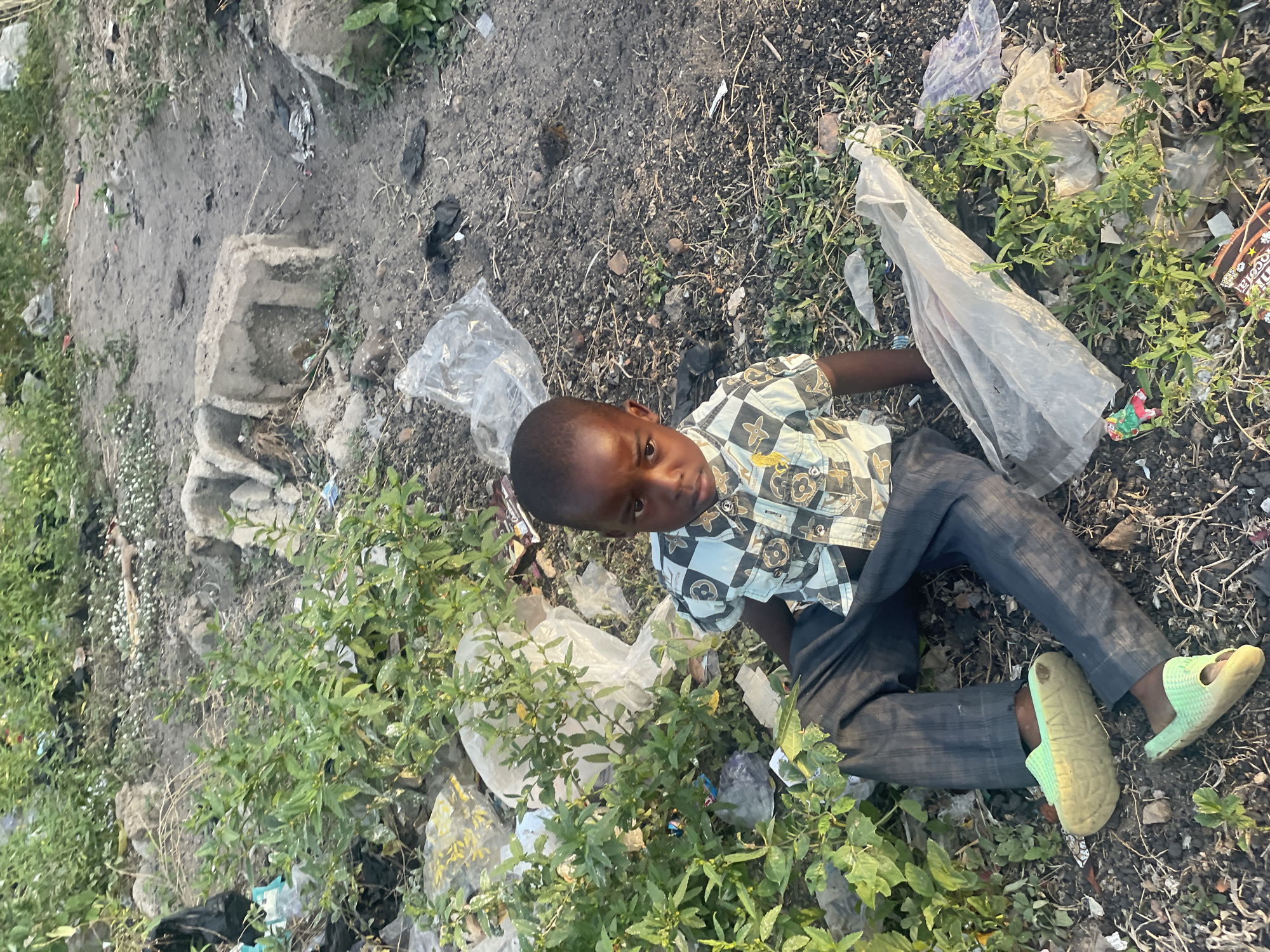 A young boy lies among debris and foliage in Lokoja, Nigeria. He is dressed in a checkered shirt, dark pants, and light green sandals. He is holding a partially transparent plastic bag. The background is littered with trash and overgrown with vegetation. A concrete structure is also present.