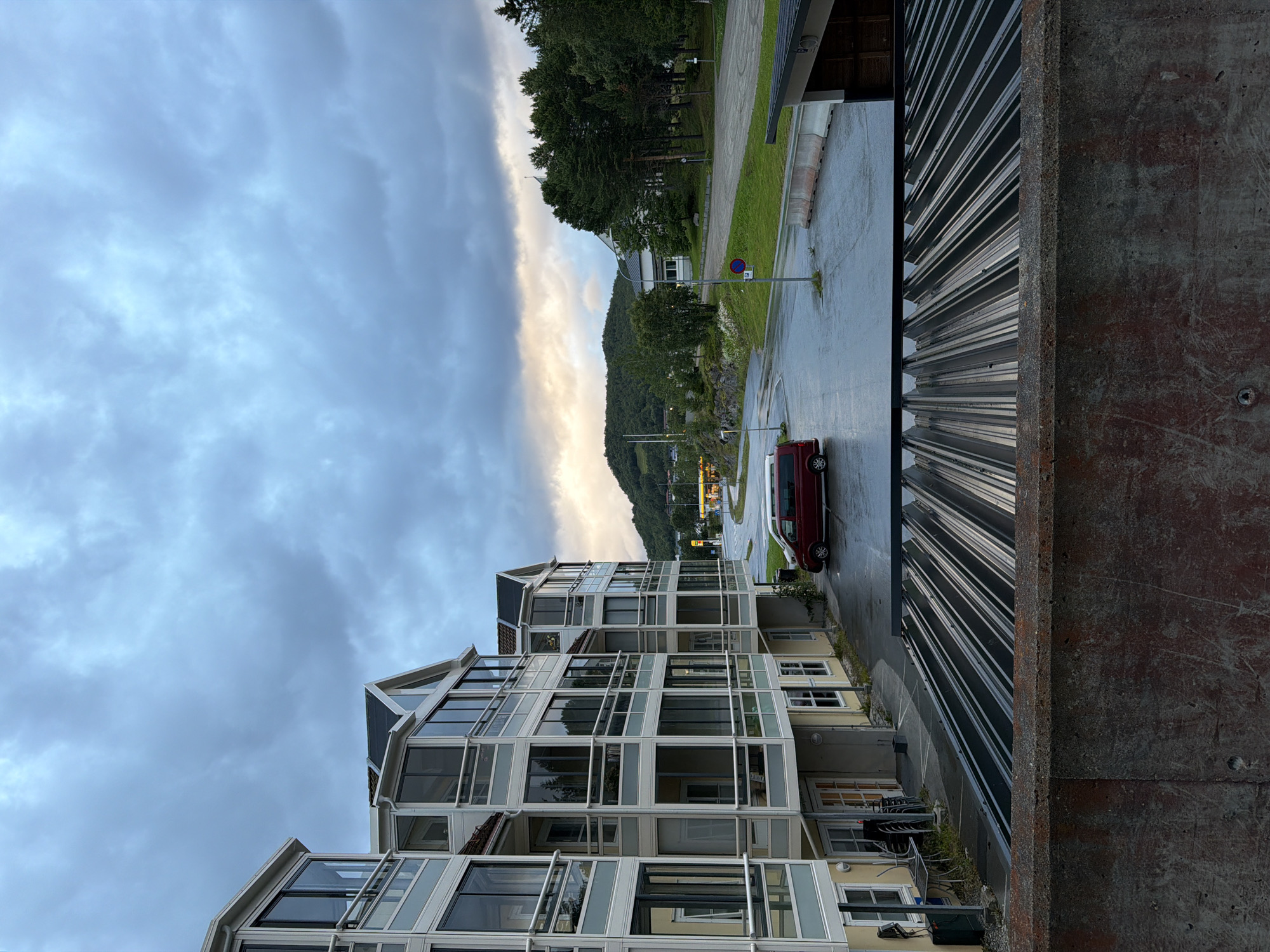 The image depicts a cloudy day scene in Harstad, Norway. In the foreground, there is a concrete structure with a rusted patina. Behind it, a corrugated metal roof is visible. The mid-ground features a multi-story apartment building with light-colored siding and extensive glass windows and balconies. A red van is parked in the wet asphalt parking lot adjacent to the building. In the background, there is a road curving to the right, leading to a gas station with yellow illuminated signs. Beyond the road, a wooded hill rises under a dramatic sky with patches of sunlight breaking through the clouds. The overall atmosphere is subdued, likely due to the overcast weather. There are no people visible in the image.