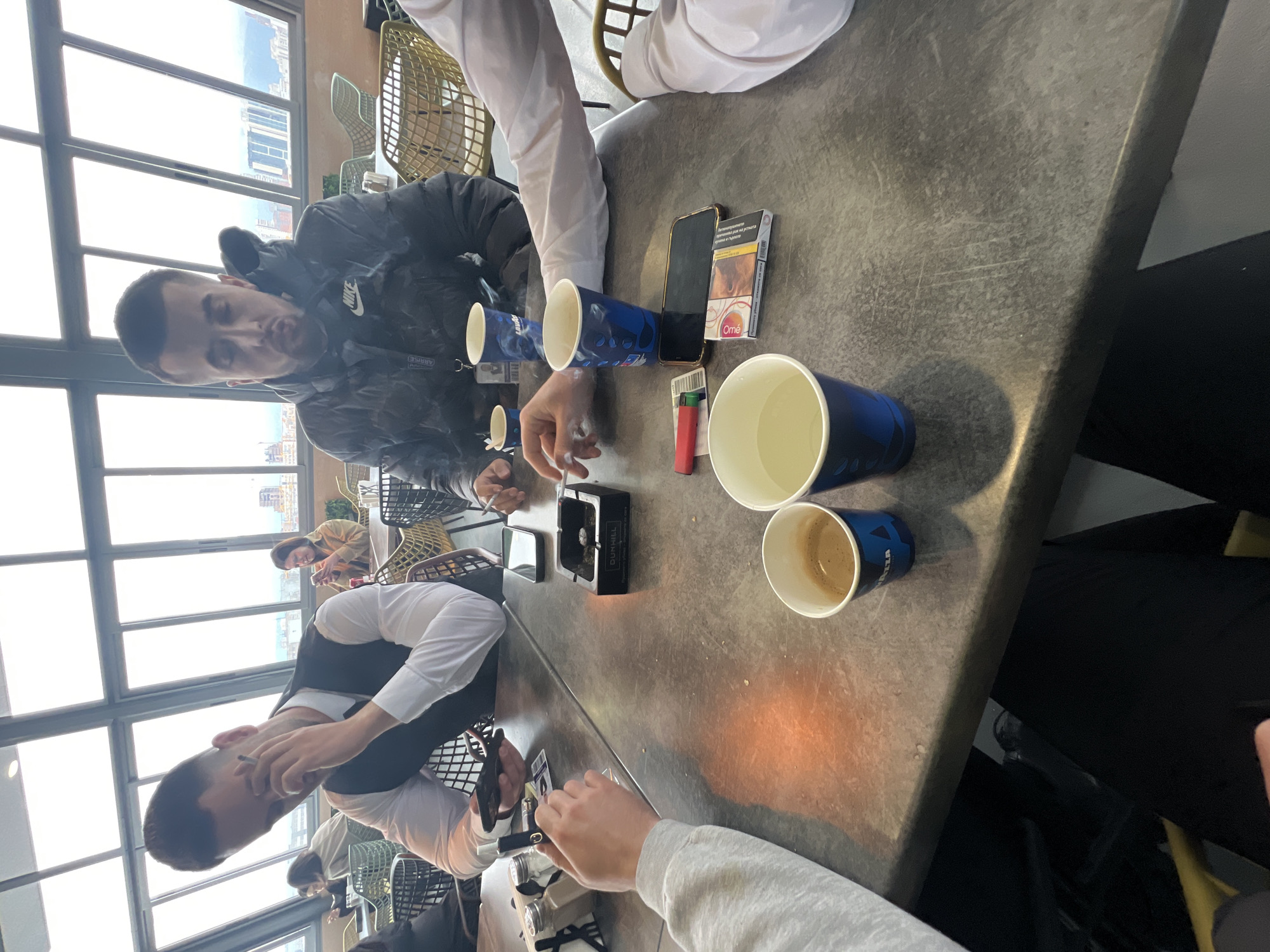 A medium, high-angle shot depicts a gray tabletop with several people sitting around it. The table contains a dark ash tray, two lighters (one red and one silver), a pack of cigarettes labeled "Omé," a black smartphone, and several blue and white paper cups, some containing coffee. People are smoking cigarettes and drinking coffee, with smoke visible in the air. The attire of the individuals includes a white long-sleeved shirt, a vest over a white shirt, and a black jacket with Nike logos. In the background, large windows reveal the cityscape of Sofia, Bulgaria.