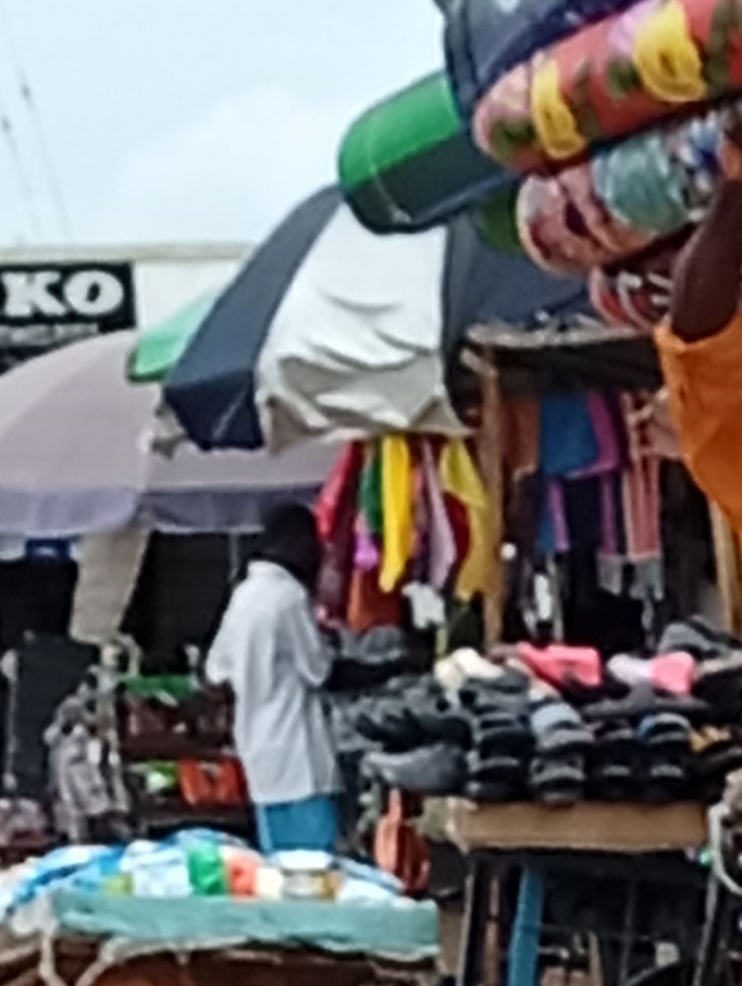 The image depicts an outdoor market scene in an unknown city. In the foreground, a table is covered with a blue and white striped cloth, displaying various items including what appear to be packaged goods and bottles. Behind the table, a person wearing a white shirt and blue shorts stands near a stall displaying colorful fabrics or scarves hanging from a structure. Another stall features a collection of shoes and sandals piled on top of a wooden surface. In the background, a large umbrella provides shade, and beyond that, a building with signage that includes the letters "KO" is visible. The overall scene suggests a bustling marketplace with various goods for sale.