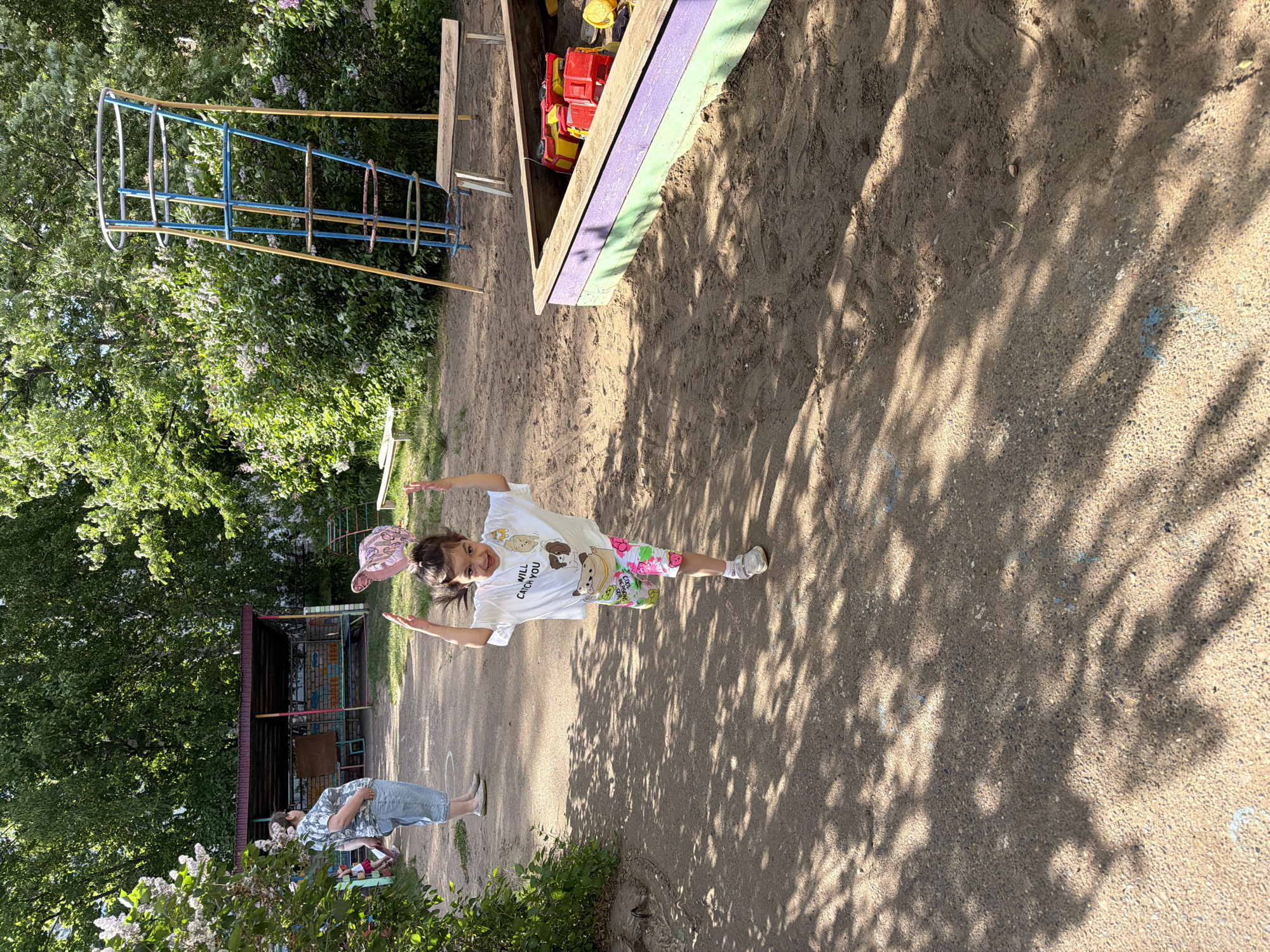 Outside in a park-like setting, a young girl wearing a white t-shirt and patterned shorts is positioned near a large, sandbox that is next to metal playground equipment. An adult is standing in the background. The ground is dirt and shows the shadows of trees. This image was taken in Chaikovsky, Russia.