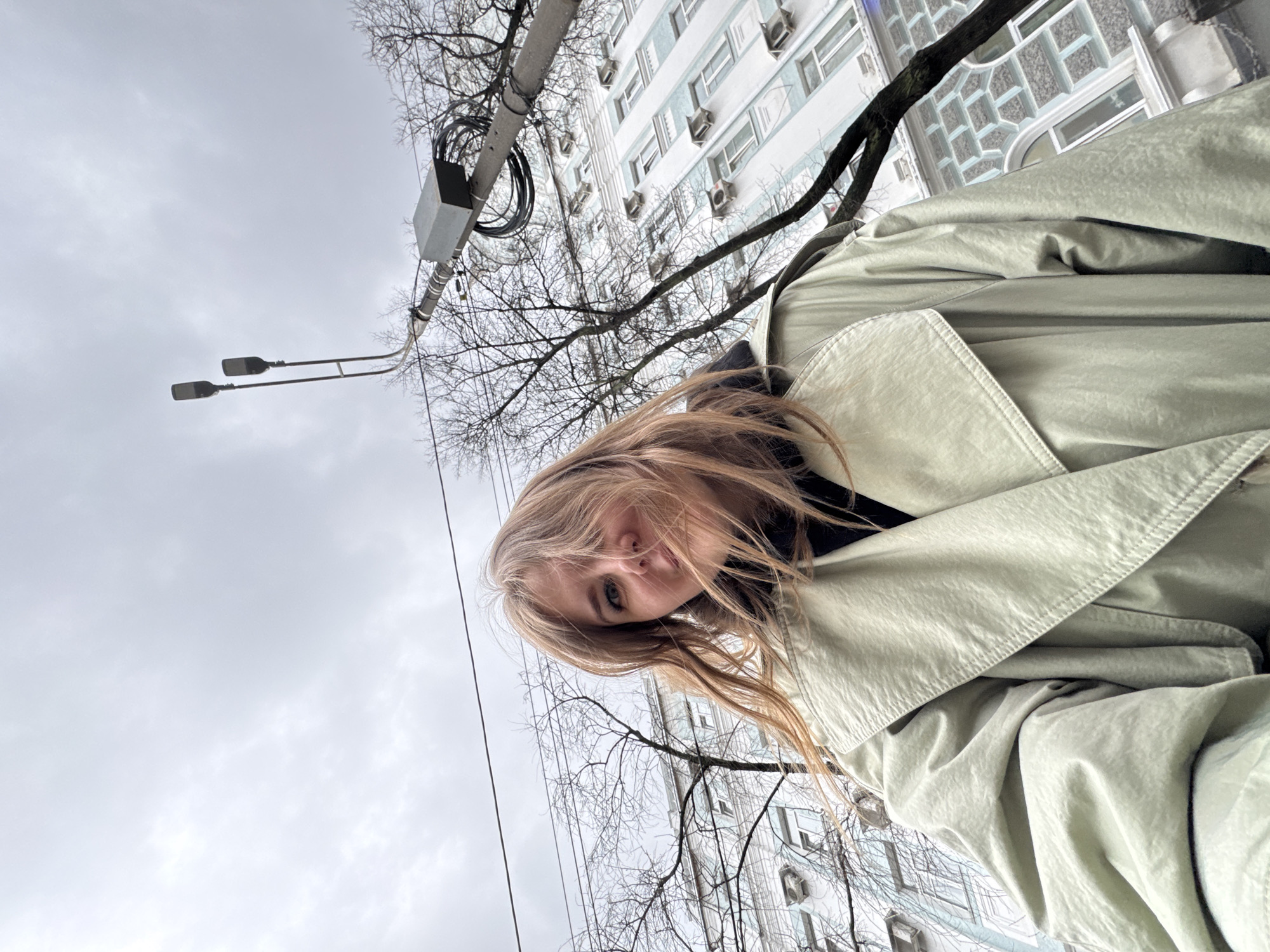 A medium-shot of a fair-skinned person with blonde hair and green eyes, wearing a light green trench coat and a black turtleneck sweater, looking up at the camera. The person is outdoors, in a city, with overcast skies above. Behind the person, there is a multi-story building with rows of windows and air conditioning units. In the foreground, there are bare tree branches, and a street lamp with two lights is visible. Based on the architecture, urban landscape, and presence of air conditioning units on the building, the location could be the city of Perm in Russia.