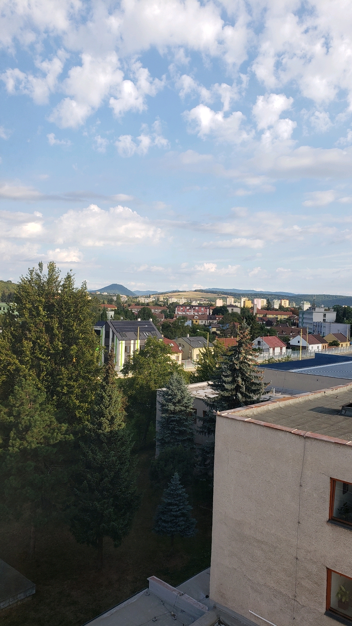 The image is a high-angle shot overlooking a town or city with buildings and trees under a partly cloudy sky. In the foreground, the corner of a tan-colored building is visible, with two windows on its side. Below and to the left of this building is a flat rooftop with some railing.
Further into the scene, dense green trees dominate the mid-ground, with some interspersed between houses. The houses are a mix of styles and colors, with some featuring pitched roofs and others flatter ones, some with solar panels.
In the background, a sprawling urban landscape with apartment buildings and smaller houses extends to the horizon. A distant, solitary hill with a rounded peak is visible on the far left. The sky is a clear blue with scattered white cumulus clouds.
This image depicts a residential area in Prešov, Slovakia, likely taken from a higher vantage point, possibly a building, looking out over the city. The time appears to be late afternoon or early evening, given the lighting and the angle of the sun, casting soft shadows. There are no people or animals visible in the frame.