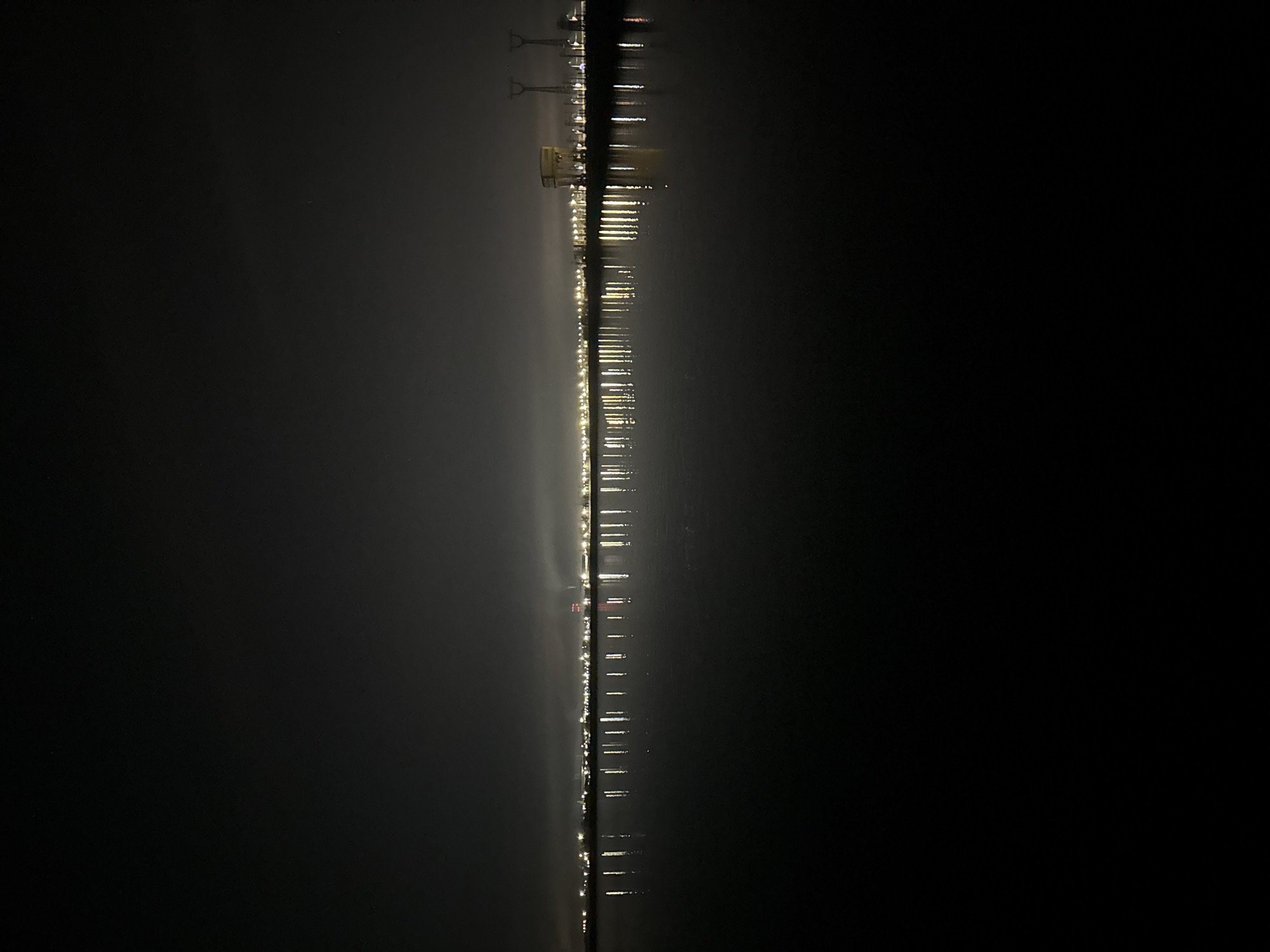 The image depicts a nighttime view of lights reflected on water, possibly a river or a large body of water. The lights are arranged in a line, suggesting a city skyline or a series of structures along the shoreline. The sky is dark and partially obscured by fog or mist, which diffuses the light. The reflections are elongated and distorted by the water's surface. The city is Volzhsky, Russia. There is no information to ascertain if the person is a perpetrator or victim.