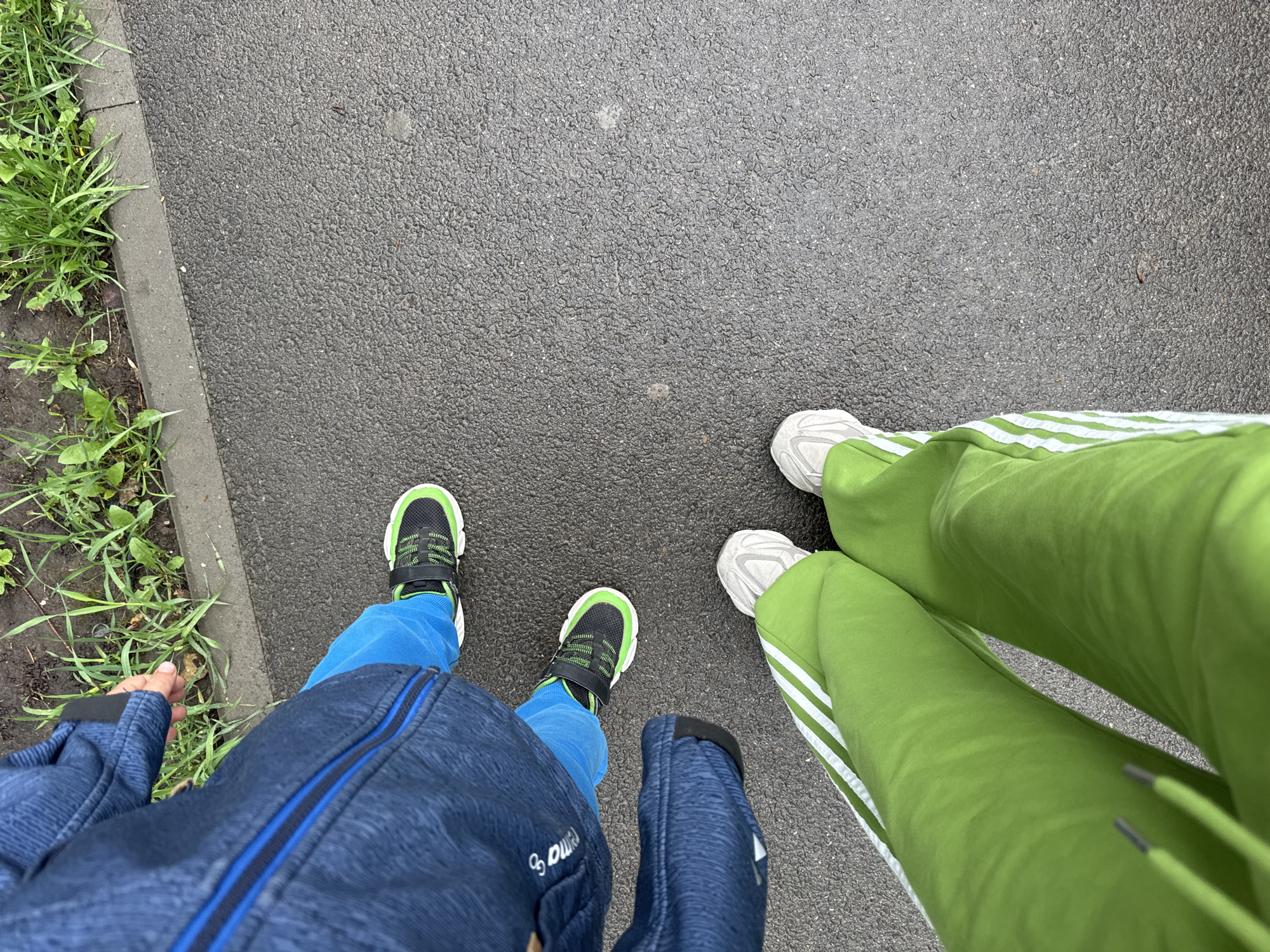 A top-down view shows two pairs of legs standing on an asphalt surface next to a patch of grass. One pair of legs is wearing blue pants with a navy-blue jacket, paired with black and green sneakers. The other pair of legs is wearing bright green sweatpants with white stripes and white sneakers. The location is Kazan, Russia.