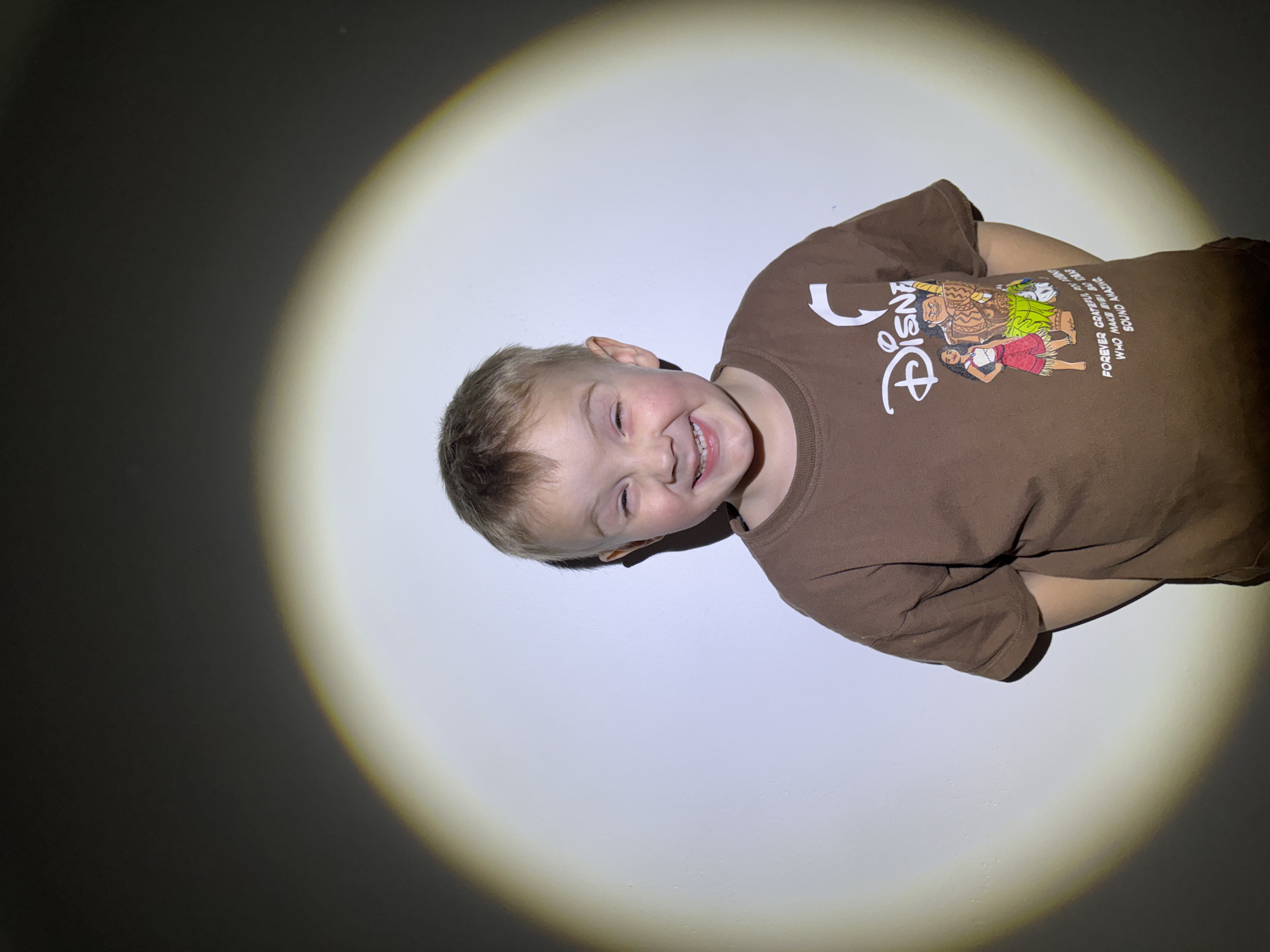 An image, rotated counter-clockwise, depicts a young boy joyfully smiling within a prominent, circular spotlight against a dark background. The boy, with light-colored hair, has a wide, happy grin, showing his teeth, and his eyes are playfully squinted. He is wearing a short-sleeved, brown t-shirt.
The t-shirt features a white "Disney" logo on the upper left side. Below and to the right of the logo, there is a colorful graphic depicting characters resembling Moana and Maui from the Disney movie "Moana." Maui is shown as a large, muscular figure with a patterned skirt, standing next to Moana, who is smaller and wearing a red skirt. Several phrases are printed in white text on the shirt, including "FOREVER WHO", "GRATEFUL IS GRACIOUS", and "SOUND AMAZING", which appear to be lyrics or quotes from the film.
The lighting effect, with a bright spotlight illuminating the boy and a dark area surrounding it, suggests an indoor setting, likely in the evening or at night. The boy's expression conveys happiness and amusement, indicating a joyful or playful moment, possibly during a photo session. The scene takes place in Chaikovsky, Russia.