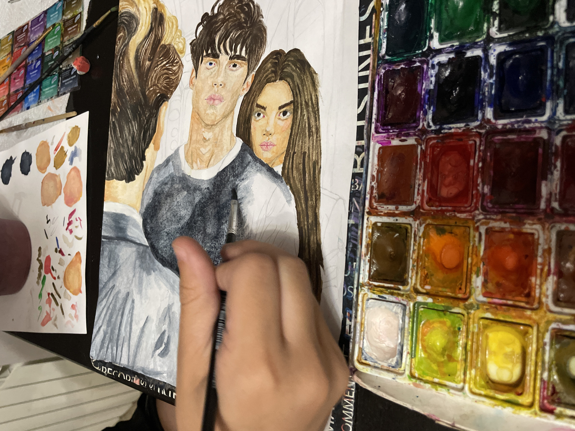 The image shows a person's hand holding a paintbrush and working on a watercolor painting of two people. The painting is laid out on a table with a watercolor palette in the foreground. The watercolor palette contains various colors, including pink, brown, yellow, orange, red, purple, blue, and green. There are also brushes and other painting supplies on the table. The background is out of focus, but appears to be a striped surface. The image does not contain any information about the location in the city of Sochi, Russia.