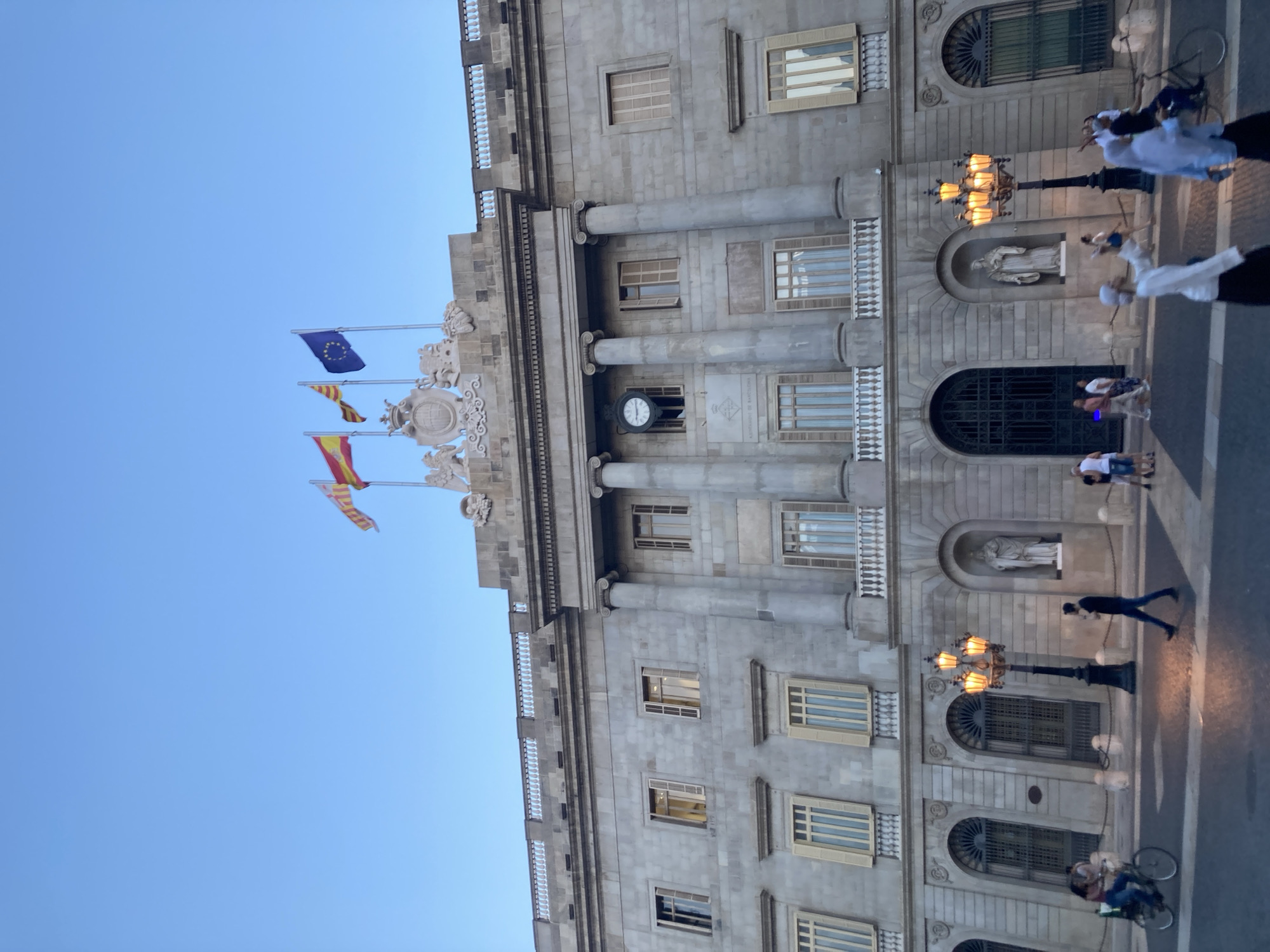 The image captures the Palau de la Generalitat de Catalunya in Barcelona, Spain, a grand stone building with classical architectural elements. Three flags, representing Spain, Catalonia, and the European Union, fly from the rooftop. The facade features a prominent clock, large arched windows, and decorative sculptures. In the foreground, pedestrians are seen walking and cycling on a paved plaza. The lighting suggests it is either early morning or late evening, with a clear blue sky overhead.