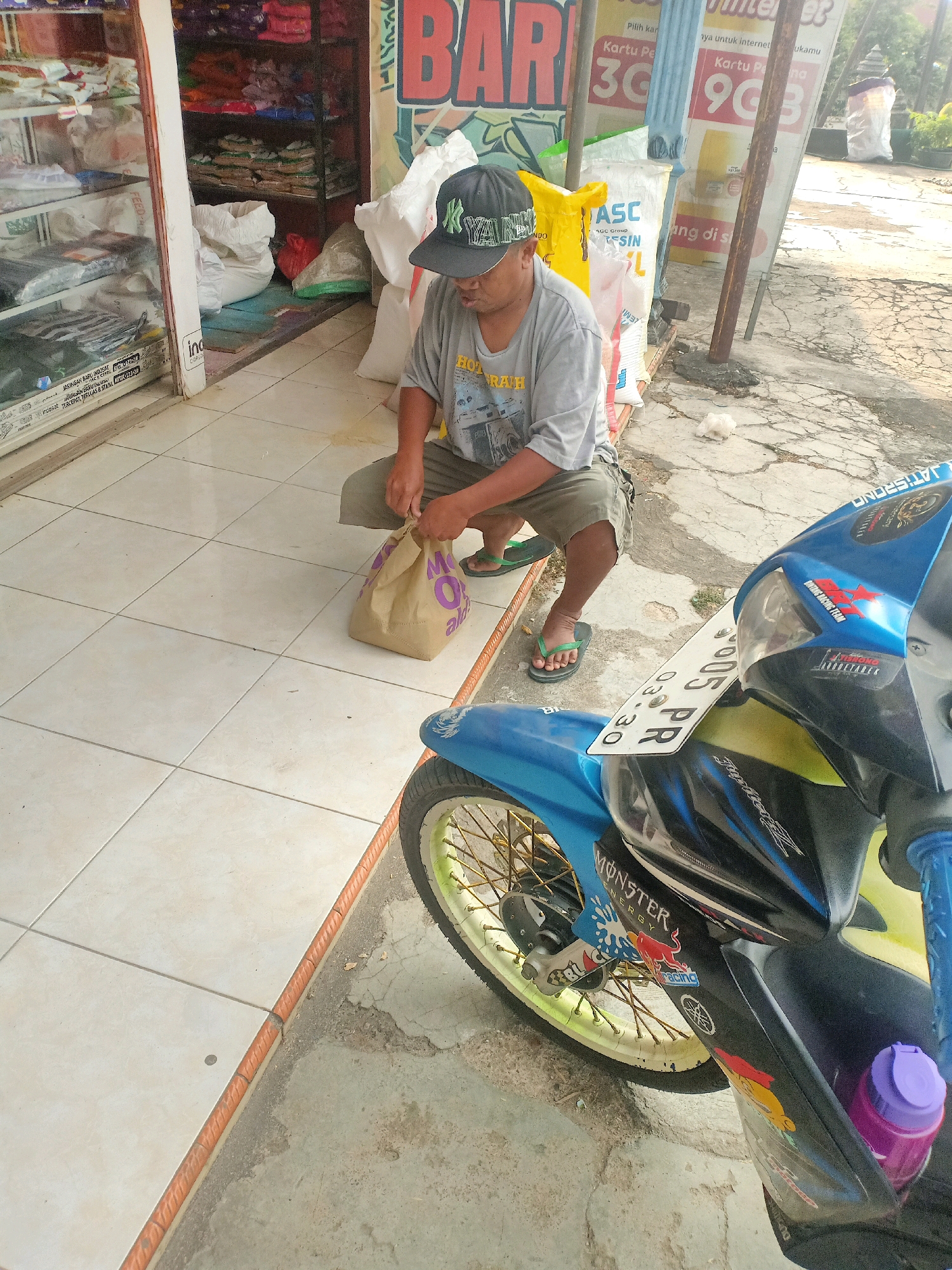 In Jatisrono, Indonesia, a person wearing a gray t-shirt, khaki shorts, and a black baseball cap kneels on a tiled floor in front of a store. The individual is holding a brown paper bag with purple lettering. To the right of the person is a blue and black motorcycle with yellow spokes on the front wheel. The background shows shelves stocked with goods inside the store and advertising posters outside. The ground is a mix of tiles and concrete.