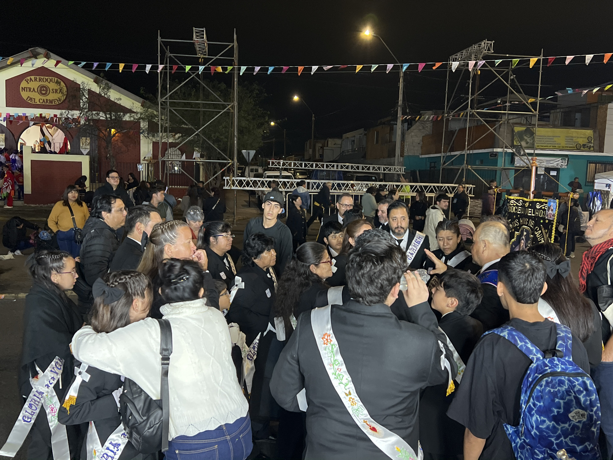This is a nighttime, eye-level, medium shot of a group of people gathered in what appears to be a religious procession or celebration in Arica, Chile. The scene is illuminated by streetlights and some artificial lighting, creating a festive yet somewhat dark atmosphere.
In the foreground, a young boy in a black suit and white sash with embroidered flowers is being embraced by a woman wearing a white cable-knit sweater. Another young girl, also in a black outfit and white sash, is standing to their left, her head bowed. Behind them, a diverse group of adults of various ages and ethnicities are visible. Many are wearing black attire with white sashes, some adorned with floral patterns and text that appears to be in Spanish, possibly indicating participation in a religious order or event.
To the left of the frame, a building with a prominent sign reading "PARROQUIA NTRA. SRA. DEL CARMEN" is visible. The building is painted in red and yellow, with arched entrances. Colorful triangular flags are strung across the scene above the gathering, adding to the celebratory mood. In the background, scaffolding and a street with houses and a streetlight can be seen.
Several individuals are looking towards the center of the gathering, where a large banner with decorative elements and text that includes "SOCIEDAD RELIGIOSA" is held aloft. Some people appear to be drinking from water bottles, suggesting the event might be physically demanding. The overall impression is one of community and shared observance.