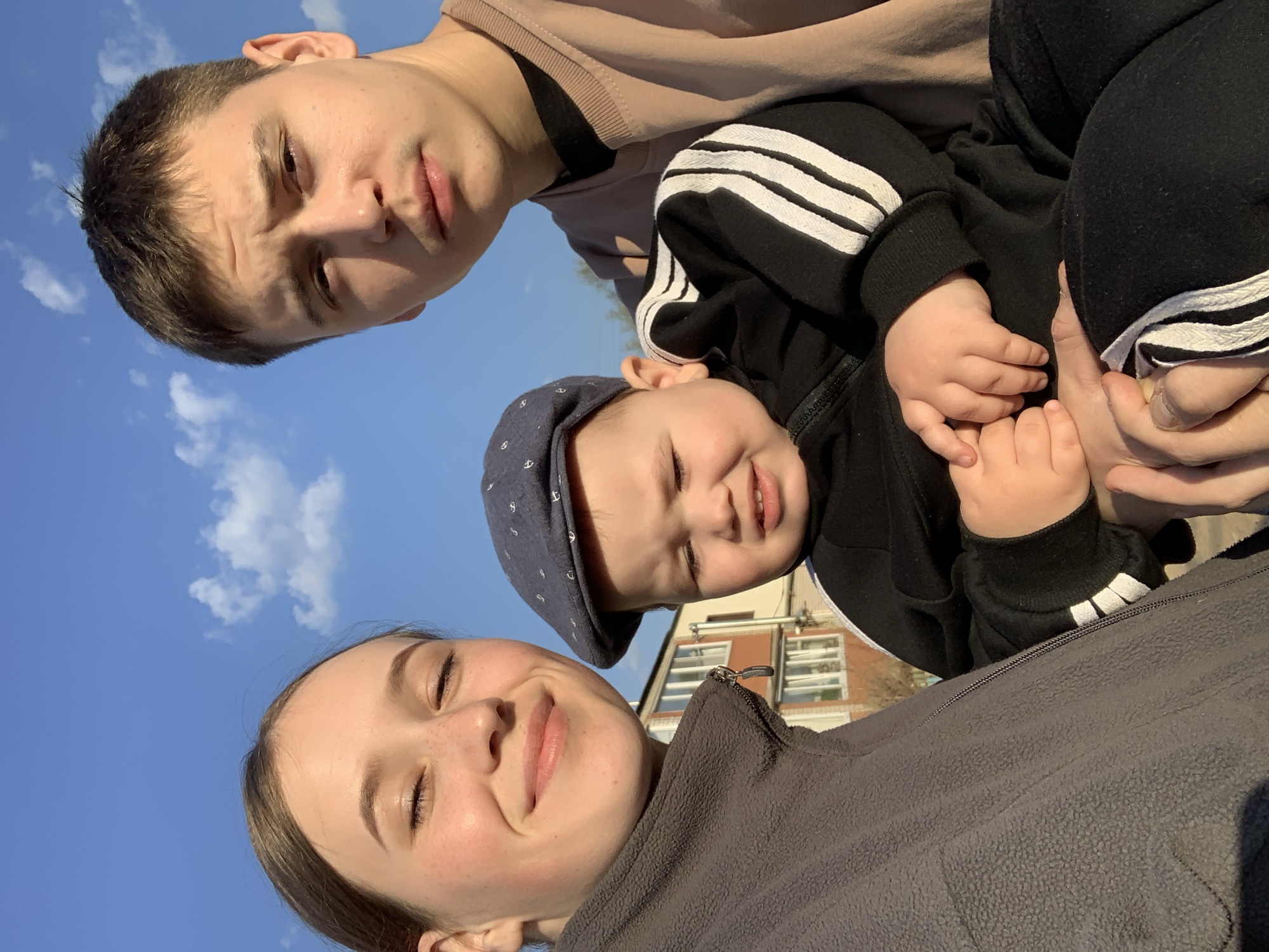 Three individuals, likely family members, pose for a selfie outdoors on a sunny day. A young woman with her eyes closed and light brown hair is at the bottom of the frame, wearing a gray fleece jacket. Above her, a toddler wearing a navy blue cap with white anchors, a black hoodie with white stripes on the sleeves, is held by someone. The child is smiling. Further up, a young man with short brown hair is visible. The background features a clear blue sky and a glimpse of a brick building with windows. The location is identified as the city of Фоки, Russia.