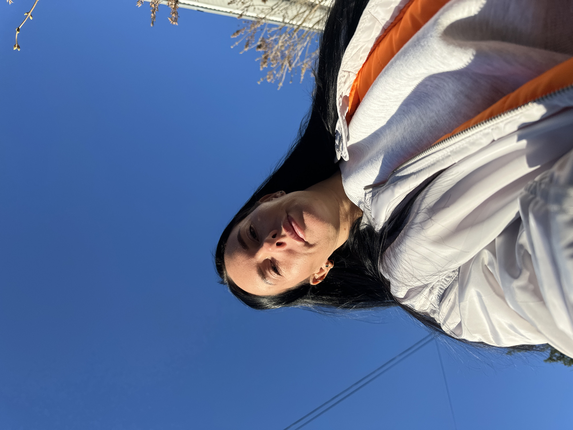 A low-angle shot shows a woman with long, dark hair wearing a white jacket with an orange lining and a grey top underneath. The background is a bright blue sky. A tree branch with buds is visible in the upper left corner. The scene appears to be outdoors. There are also cables running along the bottom left of the image. The exact location is unable to be determined.