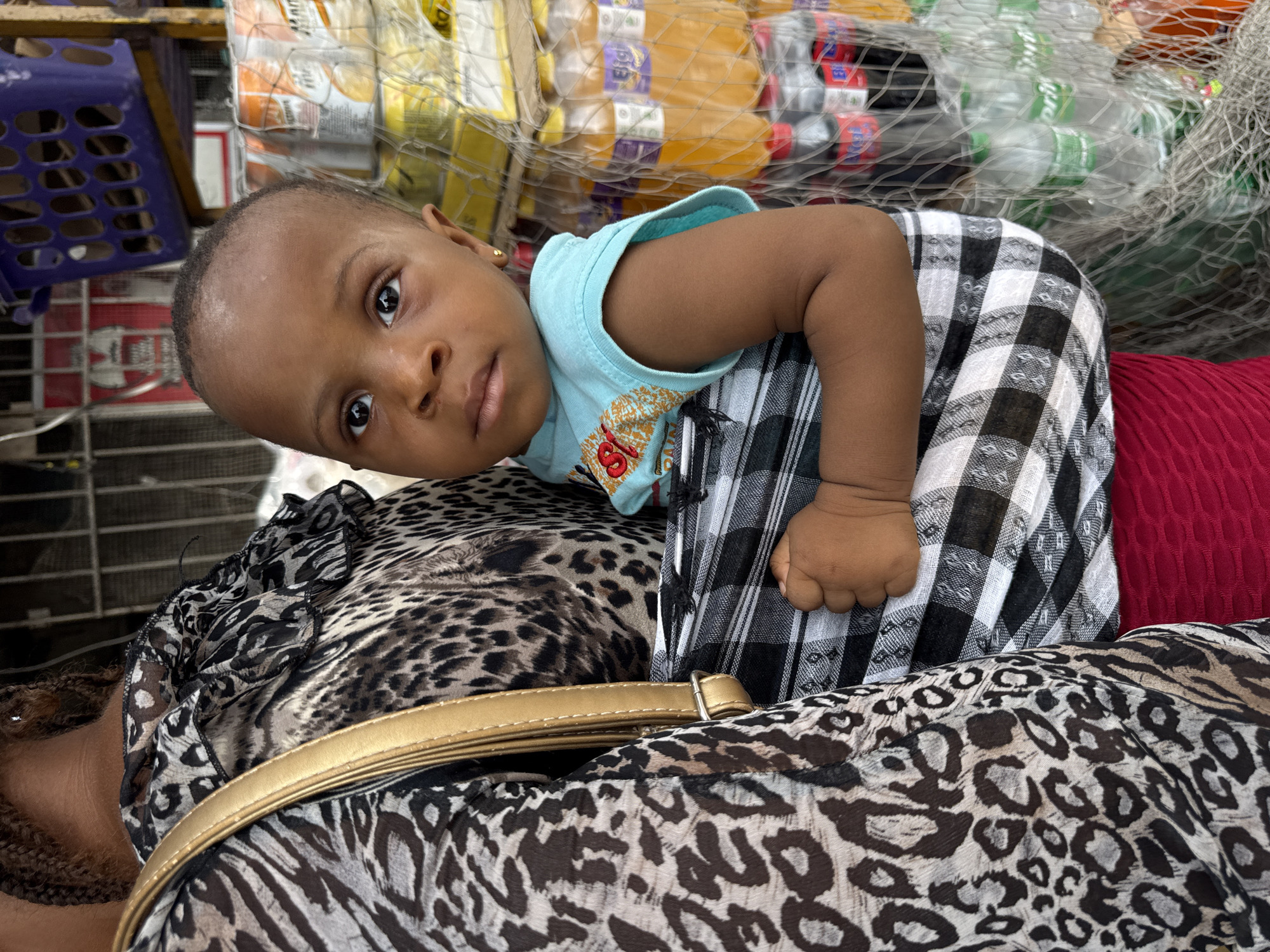 In Ado, Nigeria, a child is carried in a sling by an adult, their faces obscured. The child, wearing a light blue shirt, looks directly at the camera with a neutral expression. They are positioned in front of the adult, nestled in a black and white checkered sling. Behind them, shelves are stocked with various bottled drinks, including orange-colored beverages and dark sodas, some appearing to be in plastic netting. The background suggests a market or roadside stall environment, with glimpses of purple plastic baskets and other merchandise. The adult's attire includes a leopard-print top and a gold-colored strap is visible across their shoulder. The lighting is bright, casting a natural glow on the scene.