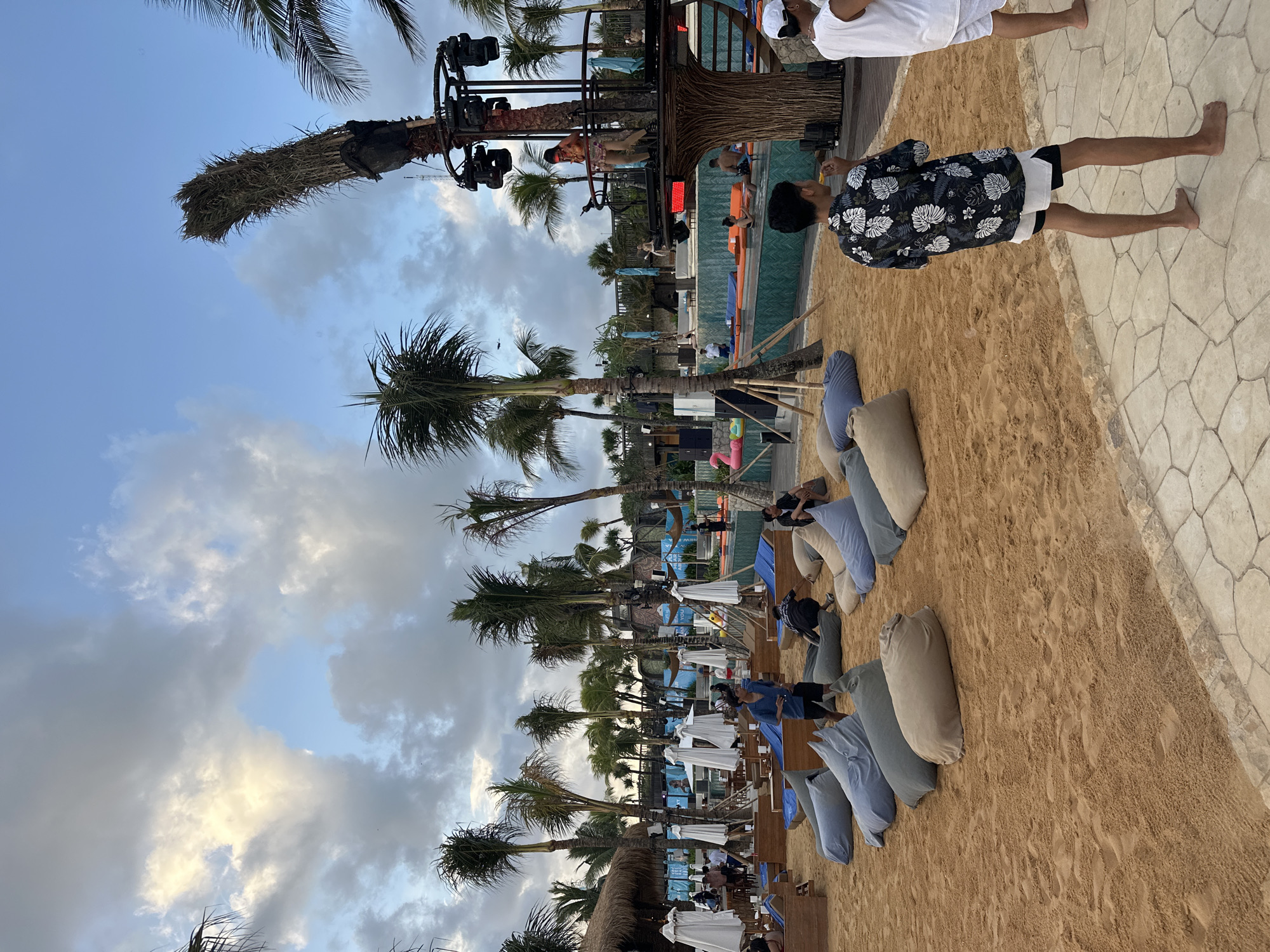 The image shows a beach setting in North Kuta, Indonesia. Palm trees line the area next to beach seating with white umbrellas. Some people are relaxing on blue and grey pillows on the sand, while others are standing on a sandy area adjacent to a paved path. In the background, there is a pool with people in and around it. Above the pool is a structure resembling a tree with a DJ booth. The sky is partly cloudy.