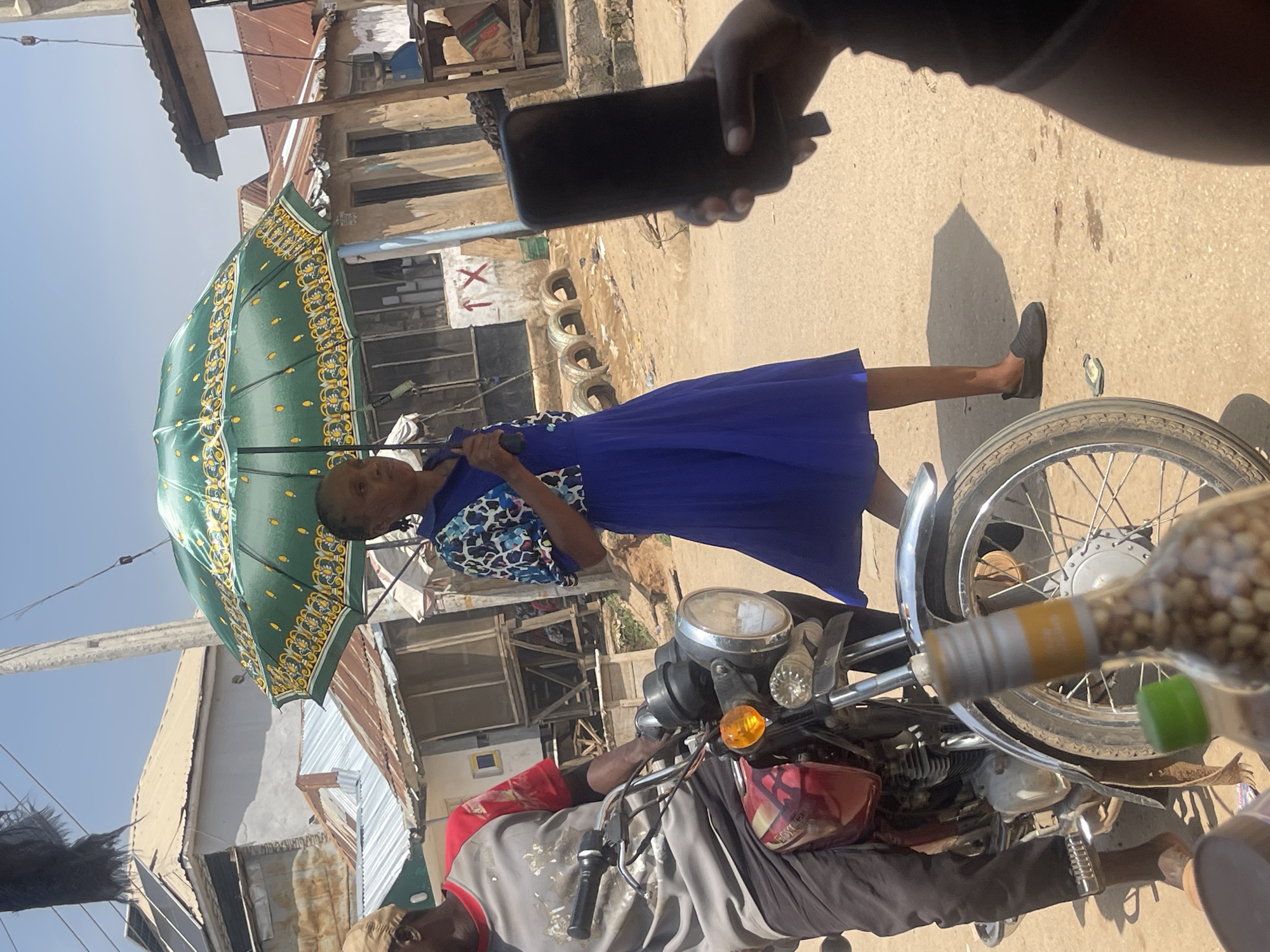 The image depicts a street scene in Abuja, Nigeria. In the foreground, a person's hand holds a dark smartphone, angled towards the camera. To the right, a motorcycle is partially visible, with its front wheel and engine prominent. A bottle, possibly containing nuts or seeds, is in the immediate foreground, blurred and out of focus.
The middle ground is dominated by two individuals. A woman, wearing a blue dress and a patterned top, is holding a large green umbrella with a decorative pattern. She appears to be walking or standing. Behind her, a person in a grey shirt with red accents is seated on or near the motorcycle.
The background reveals a bustling street with buildings, some appearing to be shops or homes with corrugated metal roofs and light-colored walls. There are also piles of tires visible, suggesting a workshop or informal business area. The ground is a paved road, appearing dusty and sun-baked. The bright sunlight suggests it is daytime, and the clear sky indicates fair weather. The overall atmosphere appears to be that of an everyday street scene in a lively urban environment. No visible text is discernable in the image.