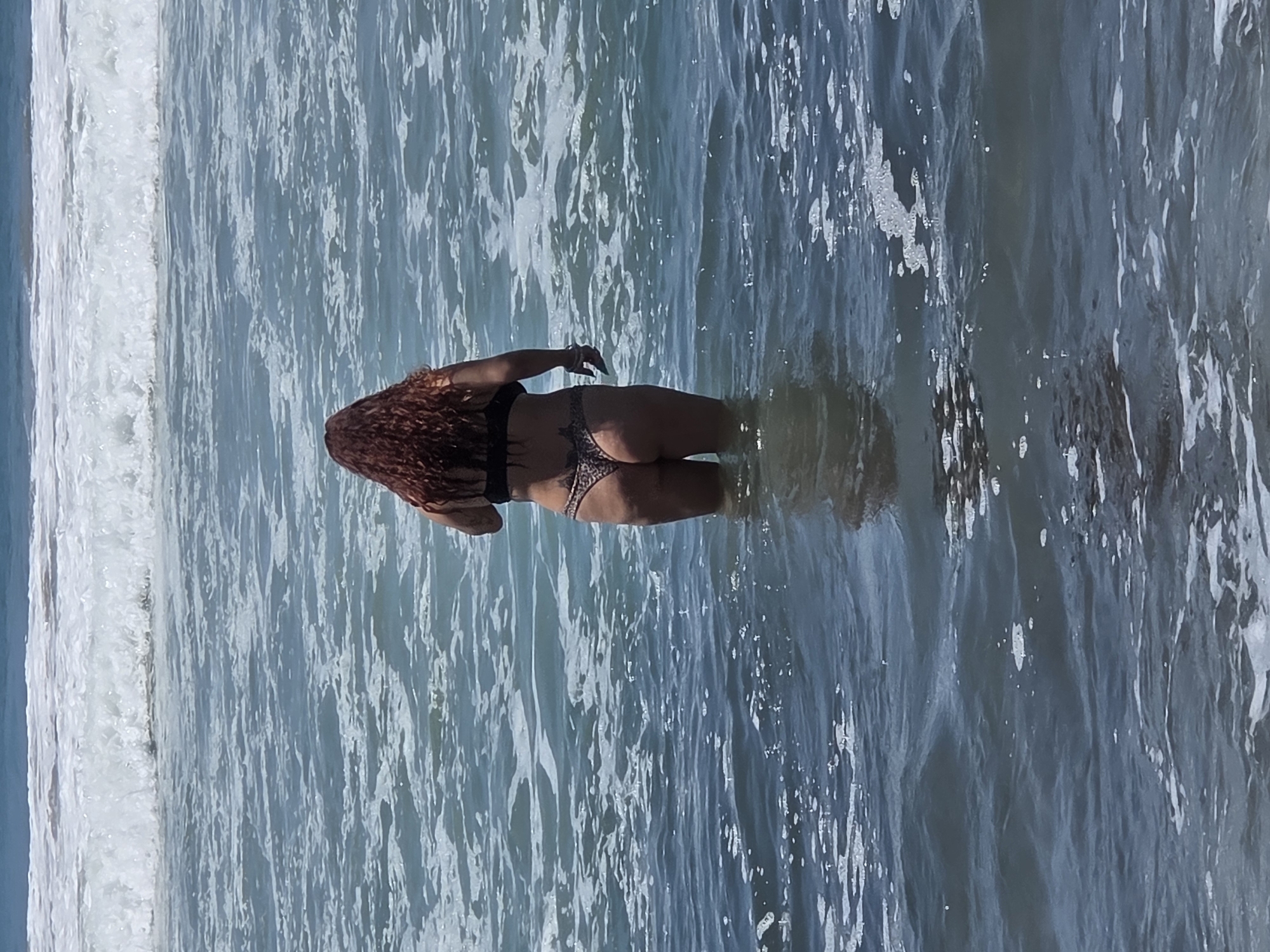 A woman with long, curly, reddish-brown hair stands in the ocean, facing away from the camera. She is wearing a black bikini with a leopard print bottom. The water comes up to her mid-thigh. In the background, there are waves breaking. The scene is set at a beach in San Clemente, California, United States of America.