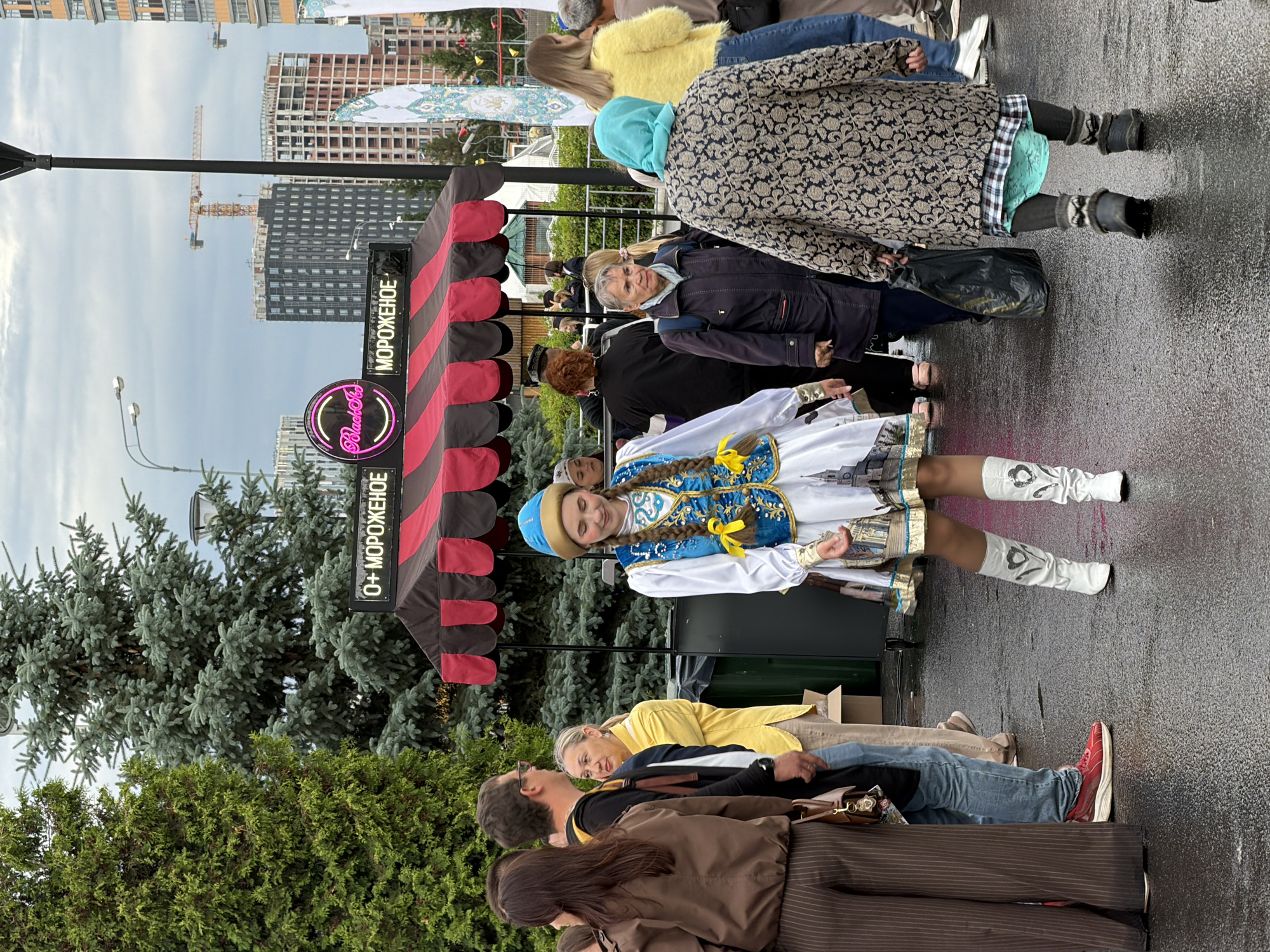 In a city setting on a wet paved surface, an outdoor event is taking place. A young woman in a white and blue traditional costume with elaborate gold embroidery and white boots is standing in the foreground, her braids falling over her shoulders. Behind her, a striped awning covers an ice cream stand with a neon sign that reads "0+ МОРОЖЕНОЕ" and "Black Star". Several people are milling about, including a woman in a patterned coat and headscarf, and a woman in a yellow shirt. In the background, tall buildings under construction and cranes are visible under a cloudy sky. The overall atmosphere appears to be casual and festive, despite the damp weather.