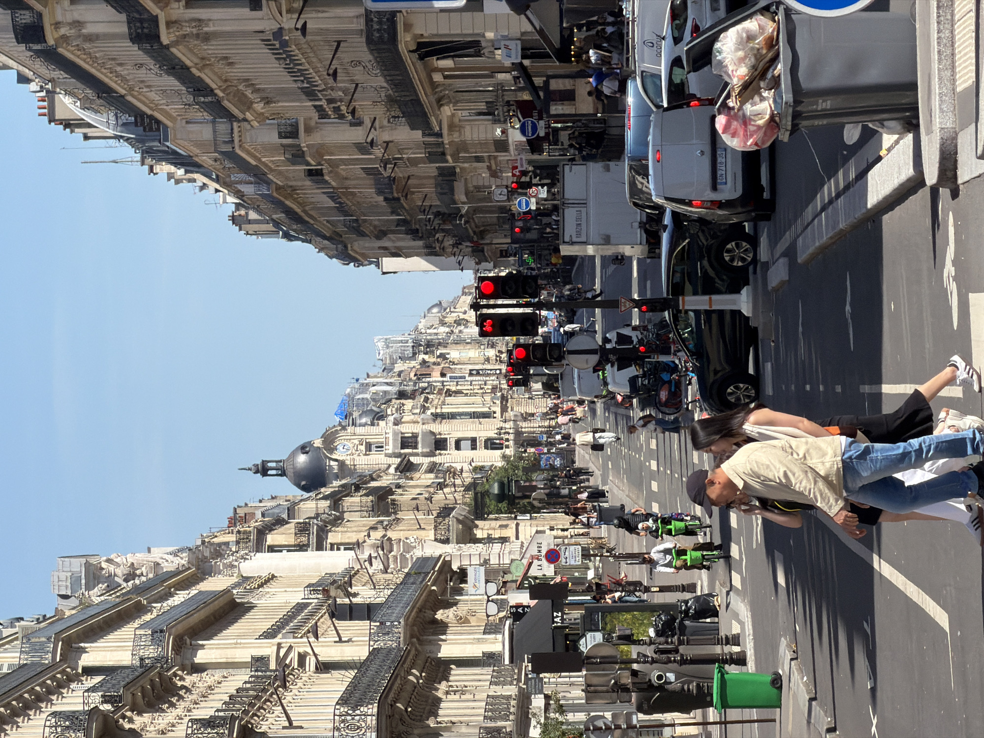 The image shows a street scene in Paris, France. The buildings lining the street feature classic Parisian architecture, with ornate facades, balconies, and mansard roofs. In the distance, a distinctive dome-topped building with a clock is visible.
The street is busy with pedestrians, scooters, and cars. Traffic lights are visible, some showing red. Several people are visible in the foreground, including a man in a tan jacket and jeans and a woman in a white tank top and black skirt. They are standing on the sidewalk. Bicycles are present along the sidewalk as well as green garbage bins.
The sky is clear and blue. The scene appears to be taken during the day, with bright sunlight.