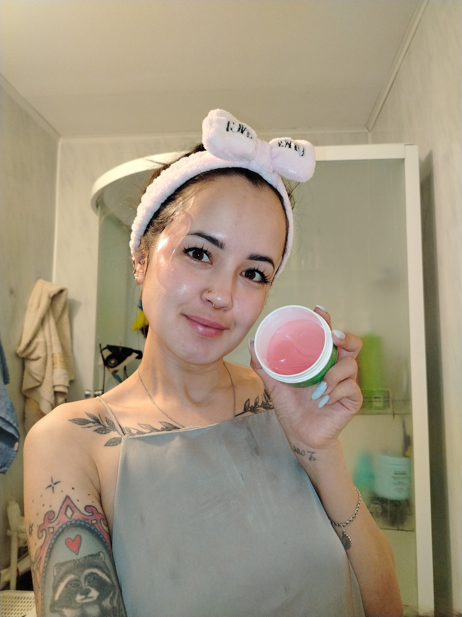 A person with light skin is posing for a picture in a bathroom. They have a pink headband with a bow on top, and eye masks under their eyes. They are holding a green and white jar with pink eye patches inside. The person has tattoos on their arm and chest. The bathroom has a shower with a glass door, and there are towels and toiletries visible. The location is Noviy, Russia.