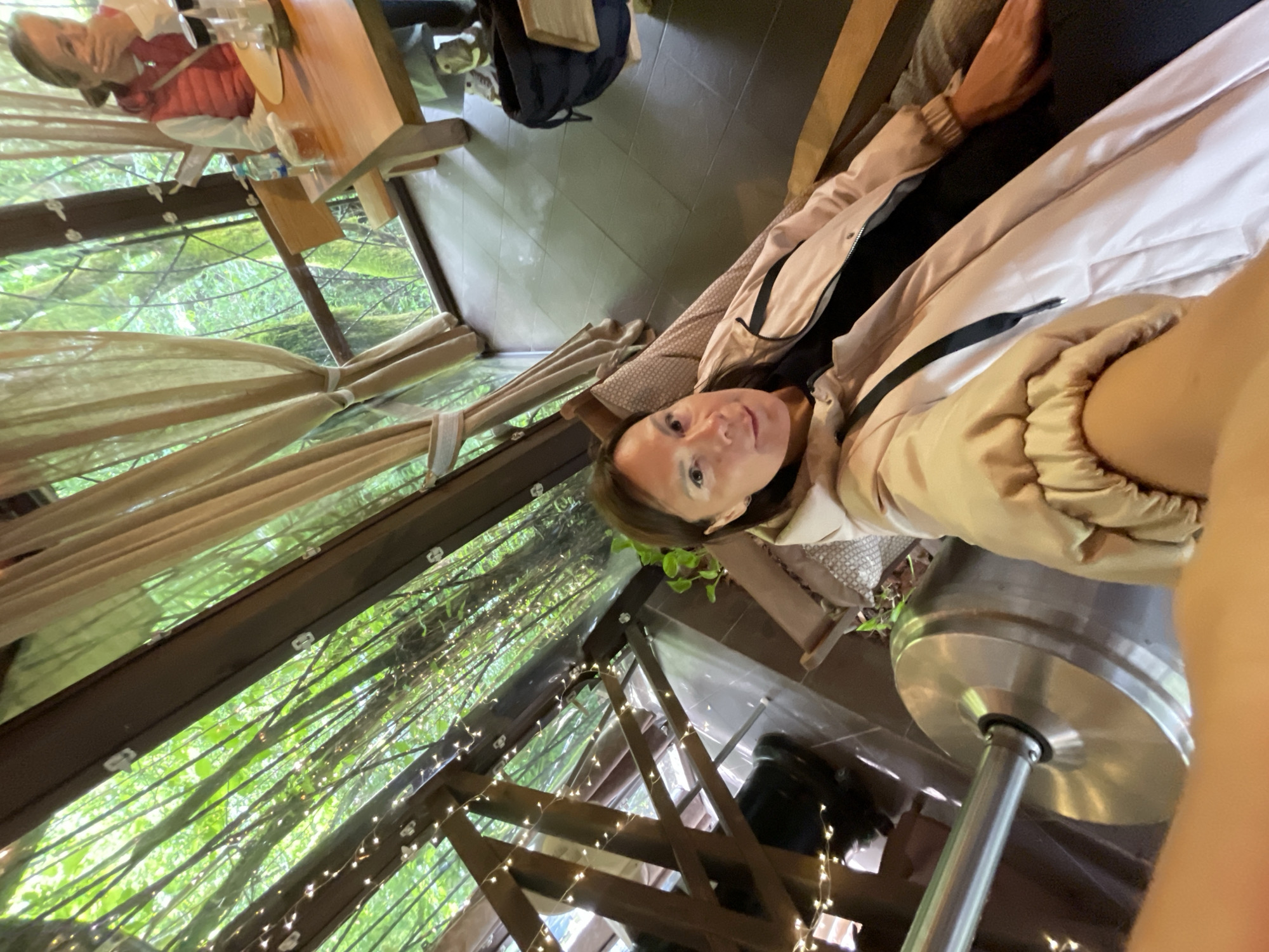The image shows a woman sitting in what appears to be a cafe or outdoor dining area, potentially in Solokh-Aul, Russia, based on provided context. She is wearing a jacket and is positioned close to the camera, possibly taking a selfie. The background includes a view of foliage or trees through a structure adorned with lights. Another person is visible at a table in the background. There is furniture and possibly a heater visible in the scene.