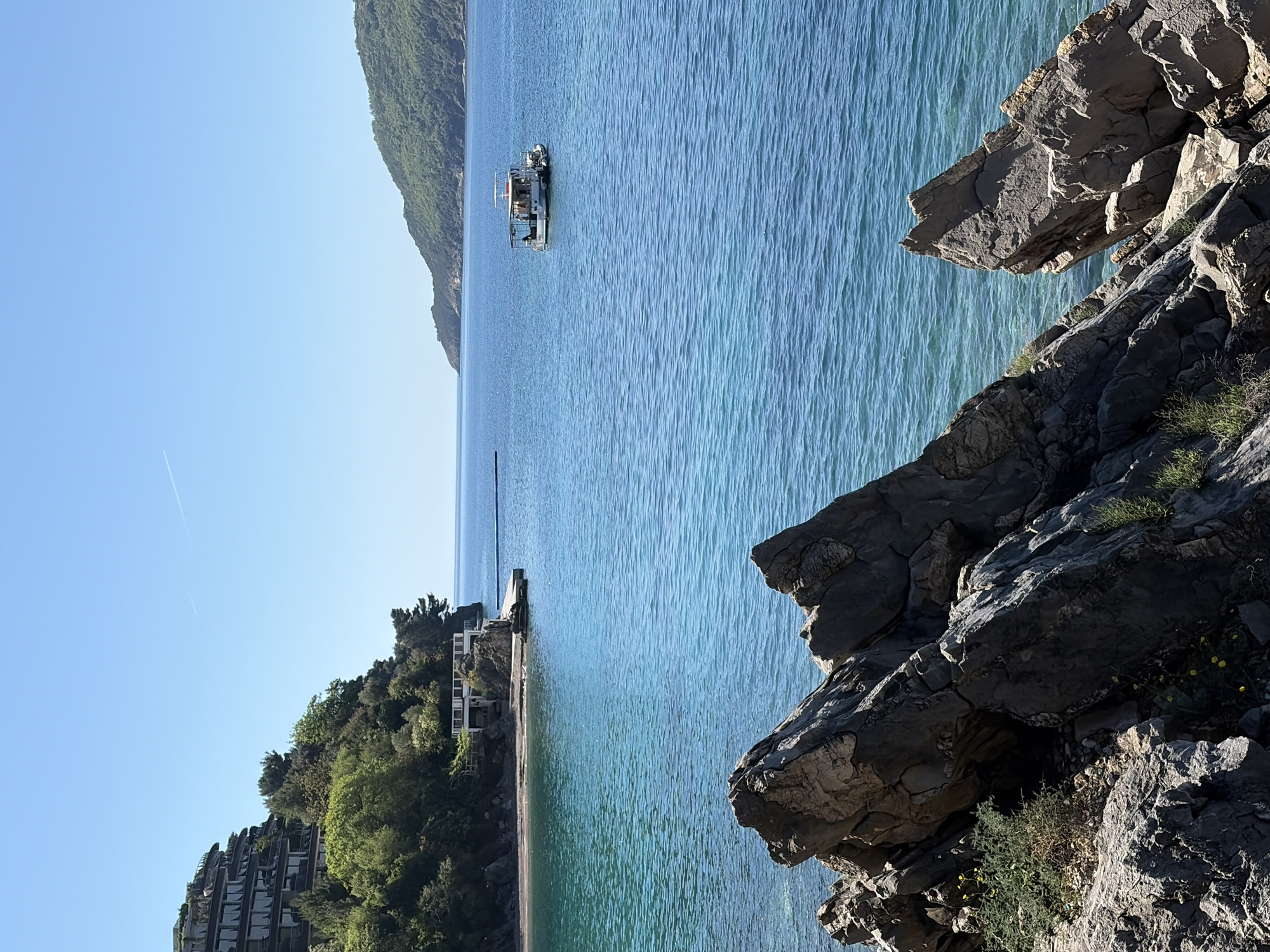 The image captures a coastal scene in Budva, Montenegro. Calm, turquoise water stretches from the foreground to the horizon. A small boat is anchored in the distance, and a long, slender pier extends into the water near a cluster of buildings nestled among green trees. Large, jagged rocks occupy the right foreground, while the edge of a hotel structure can be seen towards the lower-left corner. The sky is clear and blue.
