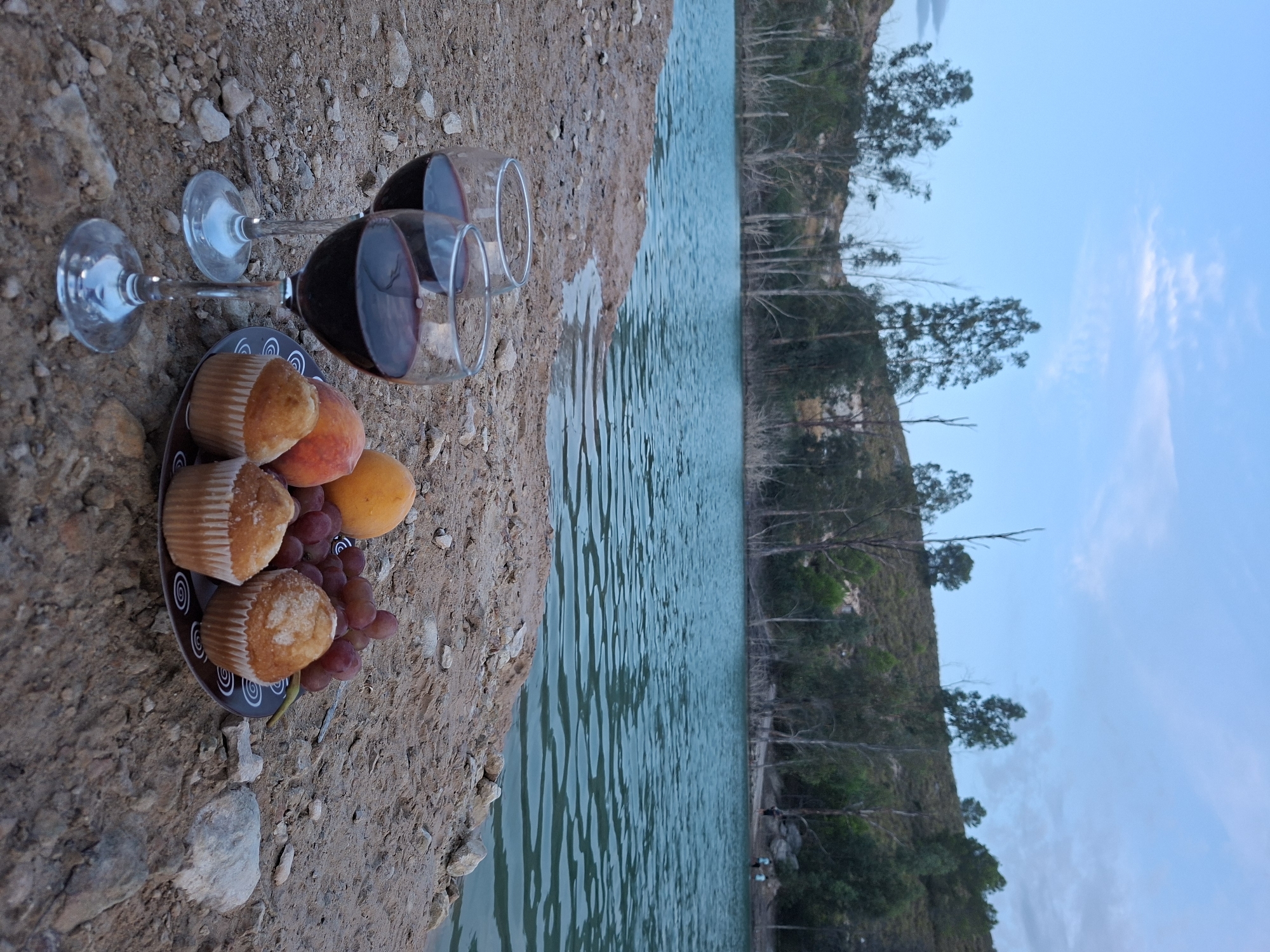 Two glasses of red wine and a plate of muffins, peaches, and grapes are set on the rocky shore of a calm, turquoise lake. In the background, a sparse line of trees separates the lake from a gently sloping, arid hillside. The sky above is a pale blue with scattered clouds. The scene suggests a peaceful, outdoor picnic. This location is likely near the Embalse de la Fuensanta, a reservoir situated in the municipality of Mula, in the Region of Murcia, Spain.