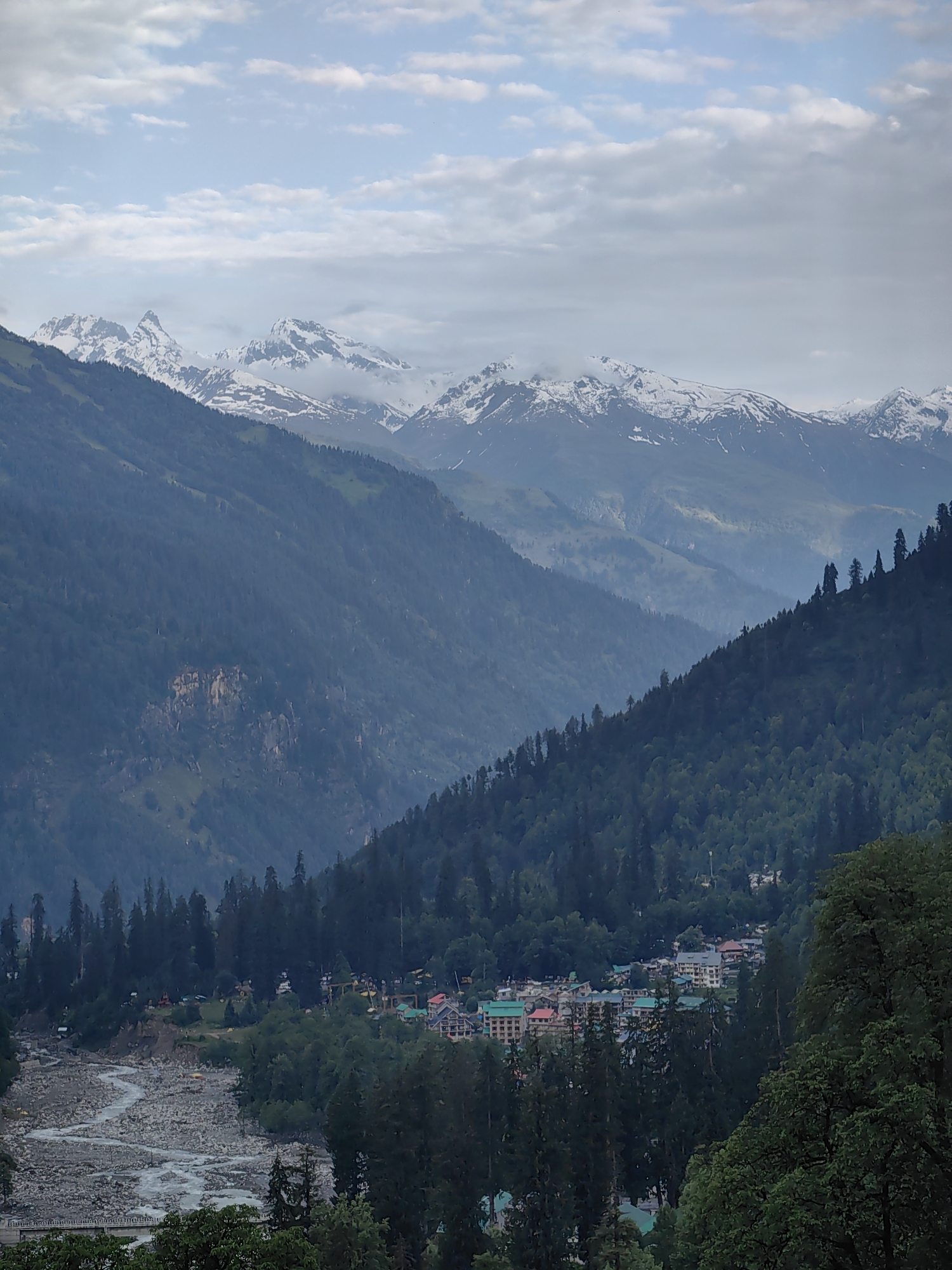 This is a vertical, eye-level, medium shot of the Solang Valley in India. The image captures a valley filled with lush green pine trees, leading to snow-capped mountains in the distance. A winding riverbed with grey stones flows through the valley, adjacent to a small settlement of buildings. The sky is overcast with white clouds, and the overall lighting suggests a subdued, hazy atmosphere, possibly due to rain or fog. No people are visible in the scene, and no actions or interactions are depicted.