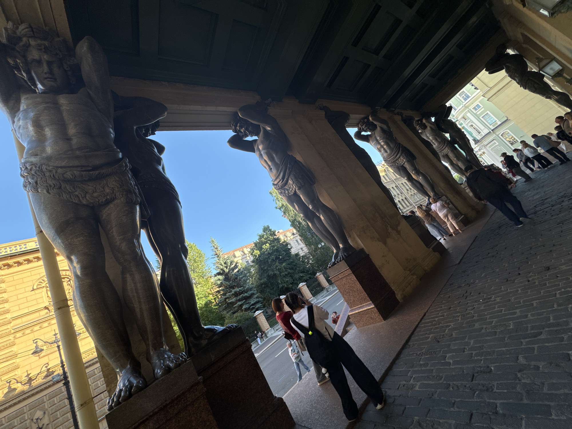 The image depicts the colonnade of the New Hermitage Museum in Saint Petersburg, Russia, featuring a row of Atlantes statues supporting the building's entablature. The perspective is low and wide-angled, looking upwards and outwards from beneath the statues. The statues are sculpted from dark stone and are depicted in various poses, with muscular torsos and legs, supporting the weight of the structure above them. In the background, a bright blue sky is visible, along with trees, buildings, and a street with pedestrians. Several individuals are walking along the street and on the pavement, some of them appearing to be tourists. One person in the foreground is facing away from the camera, wearing dark pants and a white shirt, with a backpack. The cobblestone ground is visible in the lower right portion of the image. The architectural style is classical, with ornate detailing on the building visible in the background. The lighting suggests it is daytime, with sunlight casting shadows.