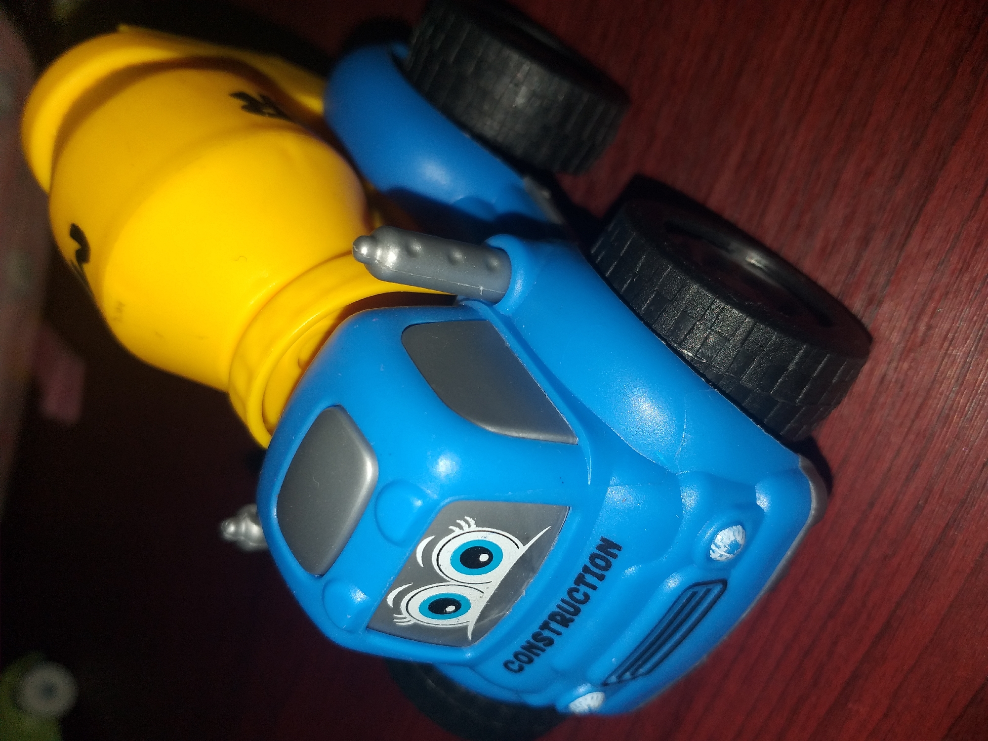 A blue toy cement truck with a yellow cement drum is shown from a low-angle perspective. The truck has cartoon-like eyes on the windshield with the word "CONSTRUCTION" printed below them. The wheels are black and appear to be made of plastic with textured treads. The truck is sitting on a dark reddish-brown surface that resembles wood grain. The background is dark and out of focus, with some blurry green and pink shapes visible. There is no indication of the location being Air Jamban, Indonesia.