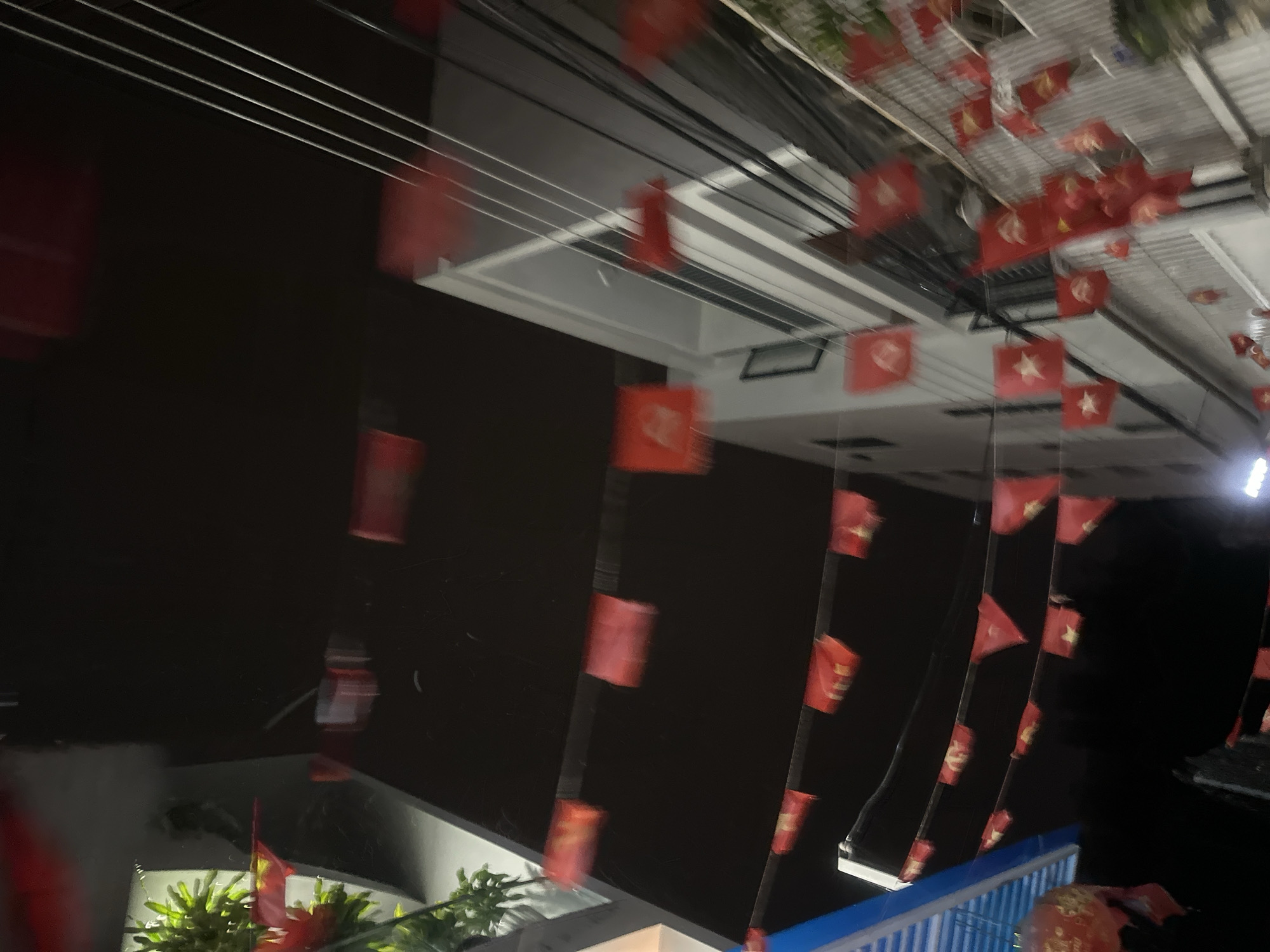 The image depicts a street scene in Bac Nha Trang Ward, Vietnam. Red flags with a yellow star emblem are strung above a street, indicating a national or political celebration. Buildings with dark exteriors and white window frames flank the sides of the street. Bright blue metal railings are visible at the bottom of the frame, possibly outlining a bridge or elevated walkway. Plants with lush green foliage are seen on the left side of the image, adding a touch of natural color to the urban setting. The lighting is subdued, suggesting the photo was taken during the evening or night.