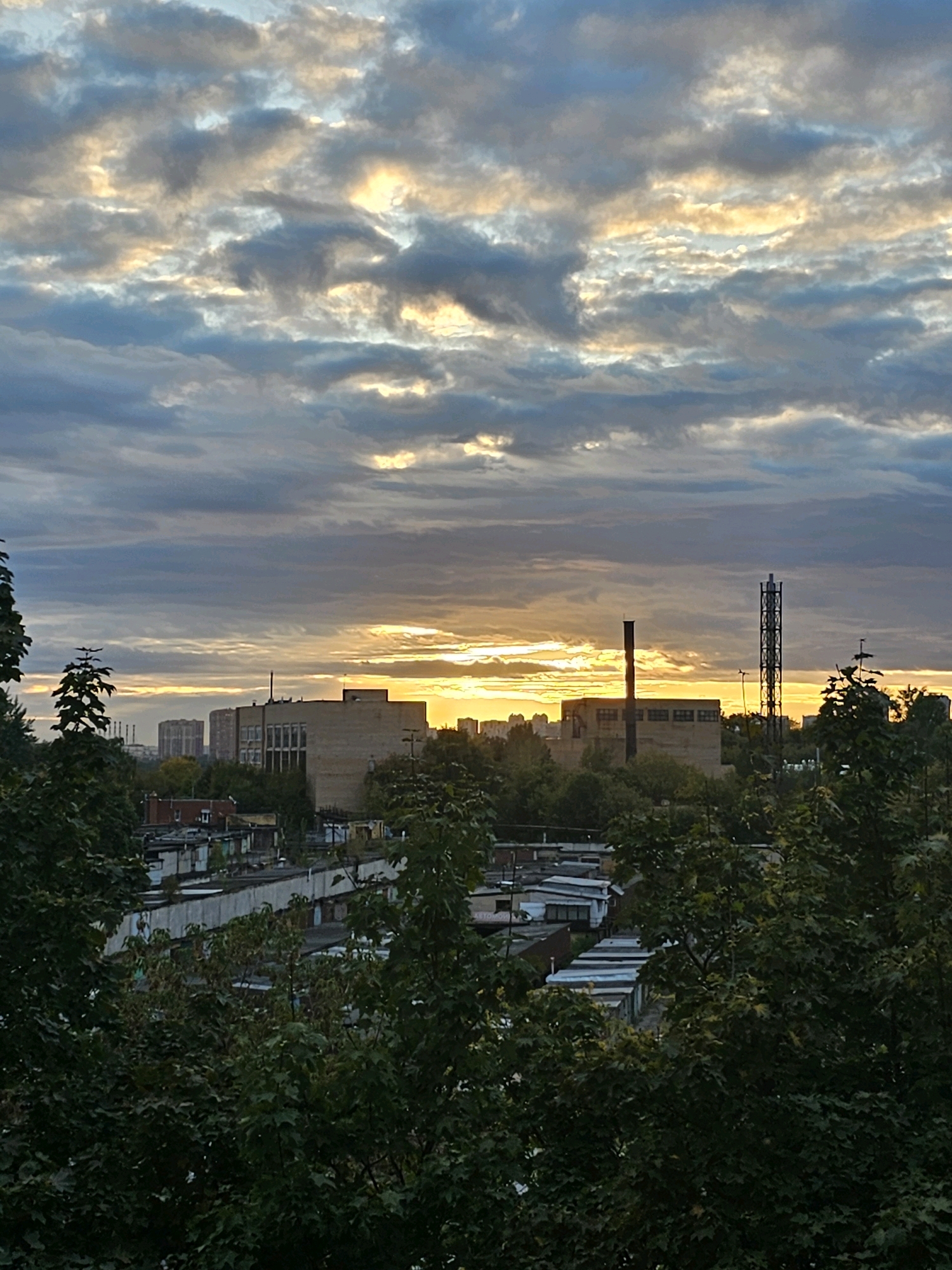 This is a vertical, eye-level, medium-shot photograph taken during sunset in the city of Khimki, Russia, capturing an industrial landscape framed by trees. The sky is filled with a dramatic display of clouds, tinged with shades of blue, grey, yellow, and orange as the sun sets. The light filters through the clouds, creating bright highlights. In the middle ground, a cluster of industrial buildings and structures is visible. One prominent building is a light-colored, rectangular structure with multiple windows. To its right, a tall, thin, black industrial tower stands with a latticed top. Further to the left, several residential buildings and what appear to be garages are arranged in rows. In the foreground, dense green trees with dark foliage occupy the lower portion of the frame, partially obscuring the view of the city below. The overall scene depicts a common urban vista where nature meets industry, illuminated by the warm glow of a setting sun.
