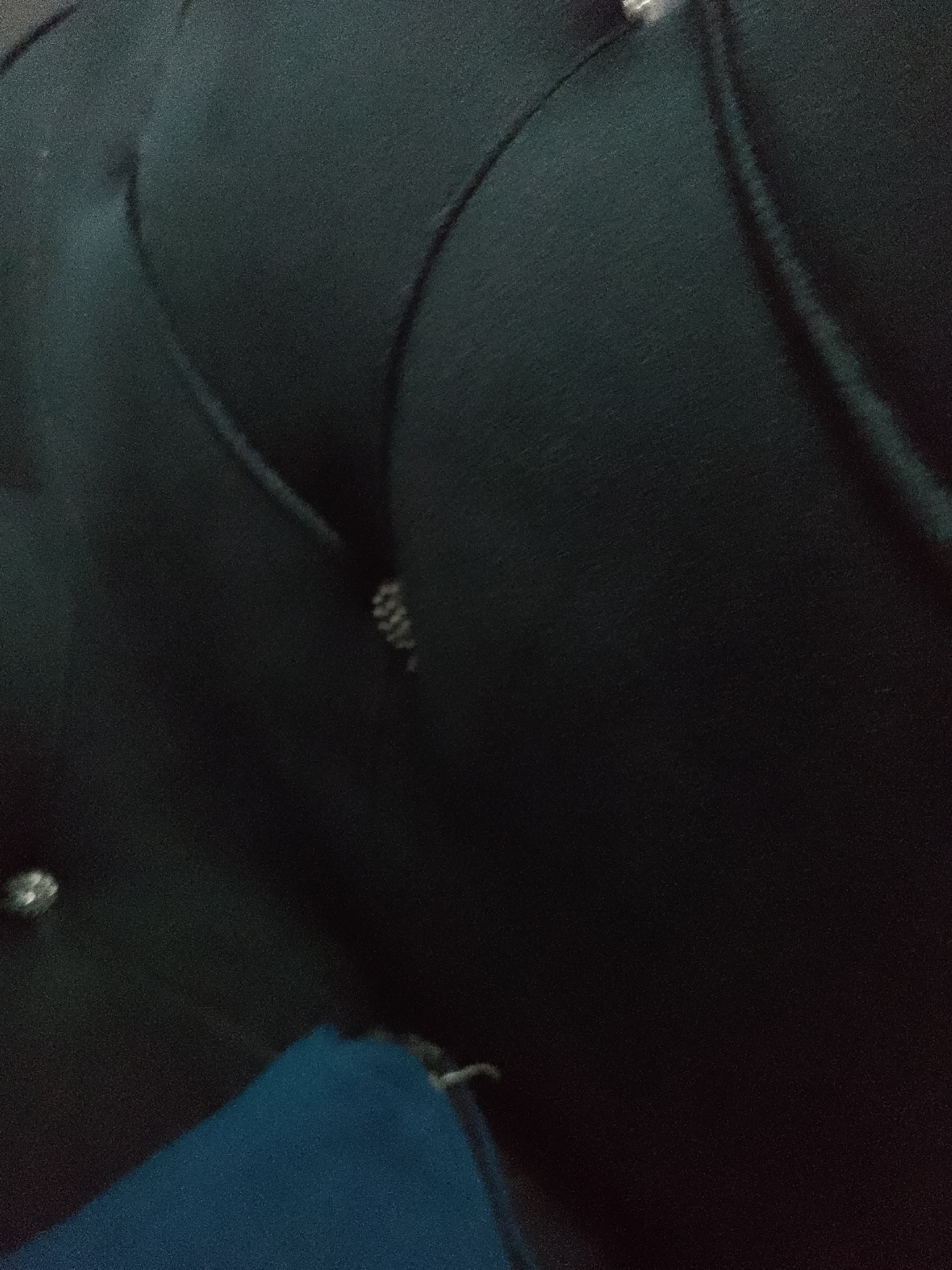The image shows a close-up of a dark, tufted surface that appears to be upholstery, possibly from a headboard or furniture. There are several buttons or studs visible on the surface, one of which has a patterned texture. A small, metallic object with a ribbed surface is also visible. In the lower left corner, a portion of a bright blue fabric is visible, partially obscuring the dark surface. The lighting is dim, creating shadows that accentuate the texture of the upholstery. There is no identifiable location in the city of Jauro Yinu, Nigeria, or any other specific geographic information in the image.